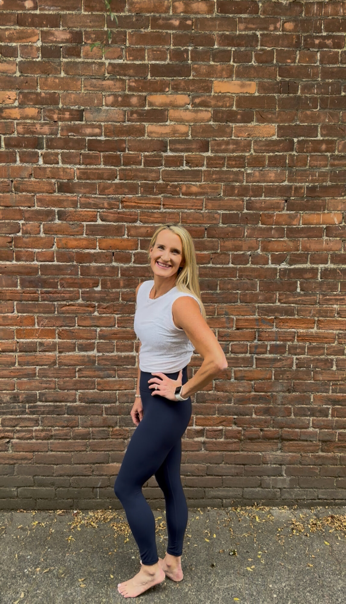 Smiling woman with blonde hair standing against a brick wall, hands on hips, wearing a white top and dark pants.