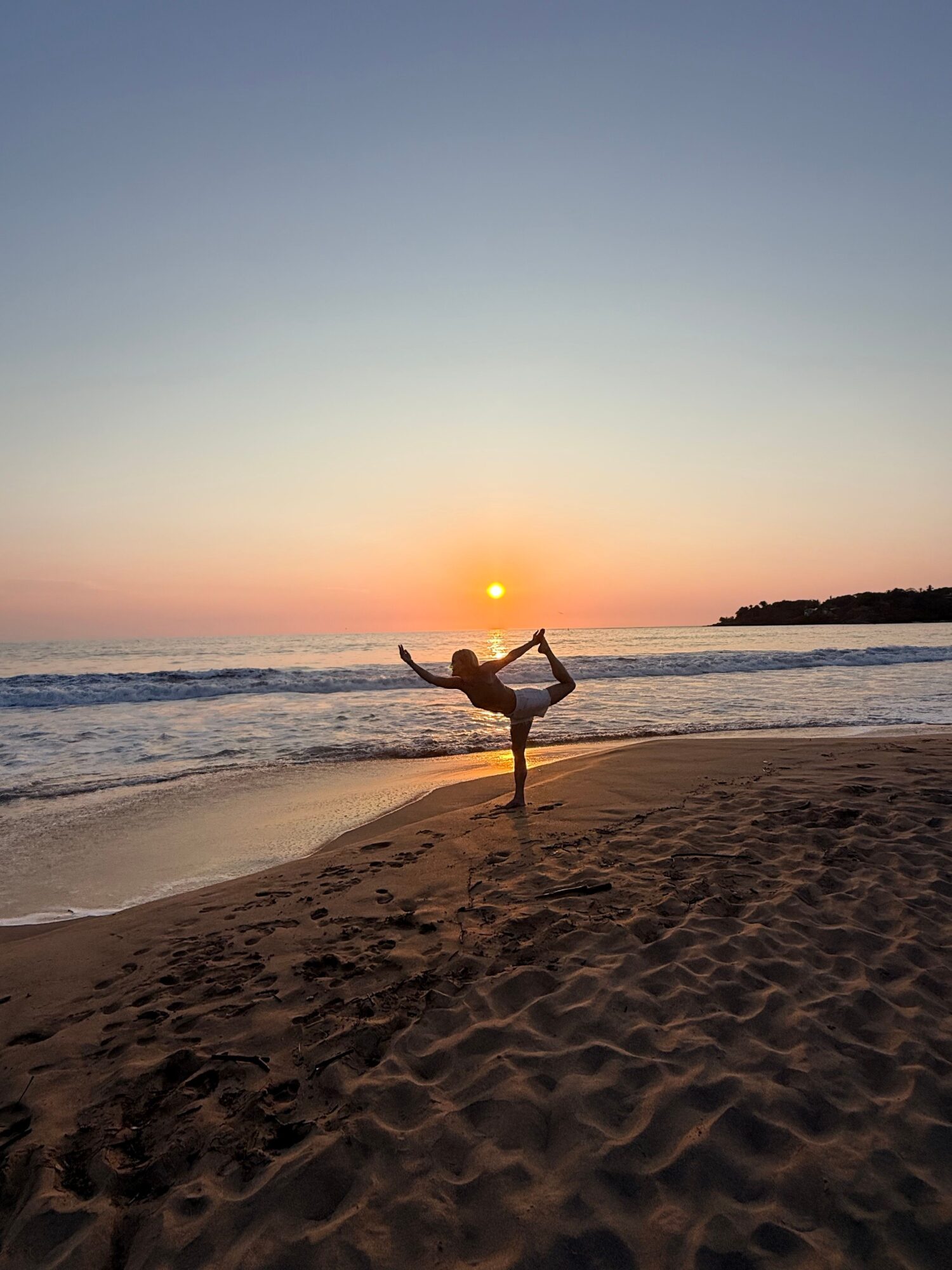 Person doing yoga on sandy beach at sunset, with ocean and colorful sky in background.