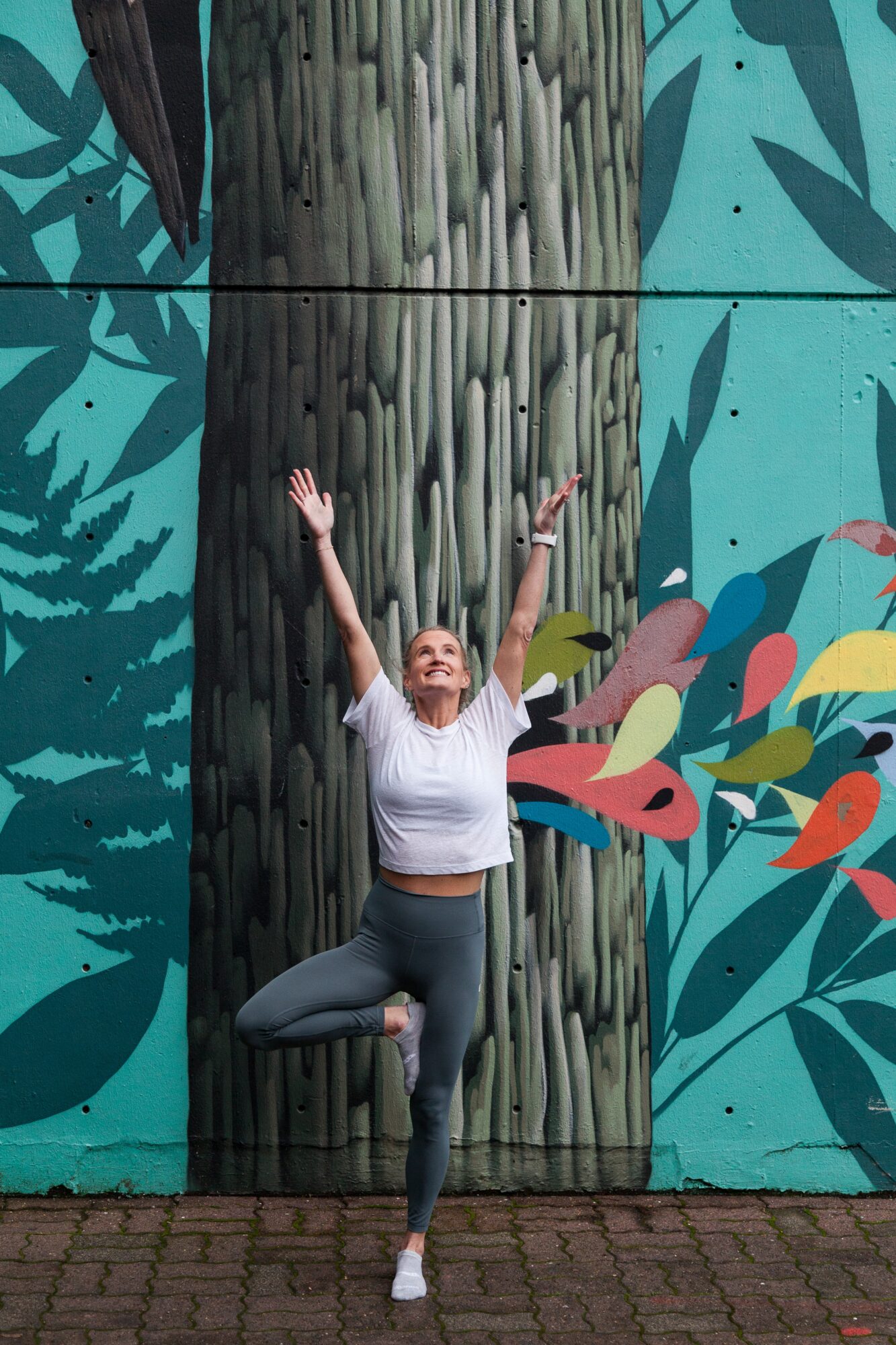 Person practicing yoga outdoors with arms raised, standing on one leg, in front of a colorful mural with plants and birds.
