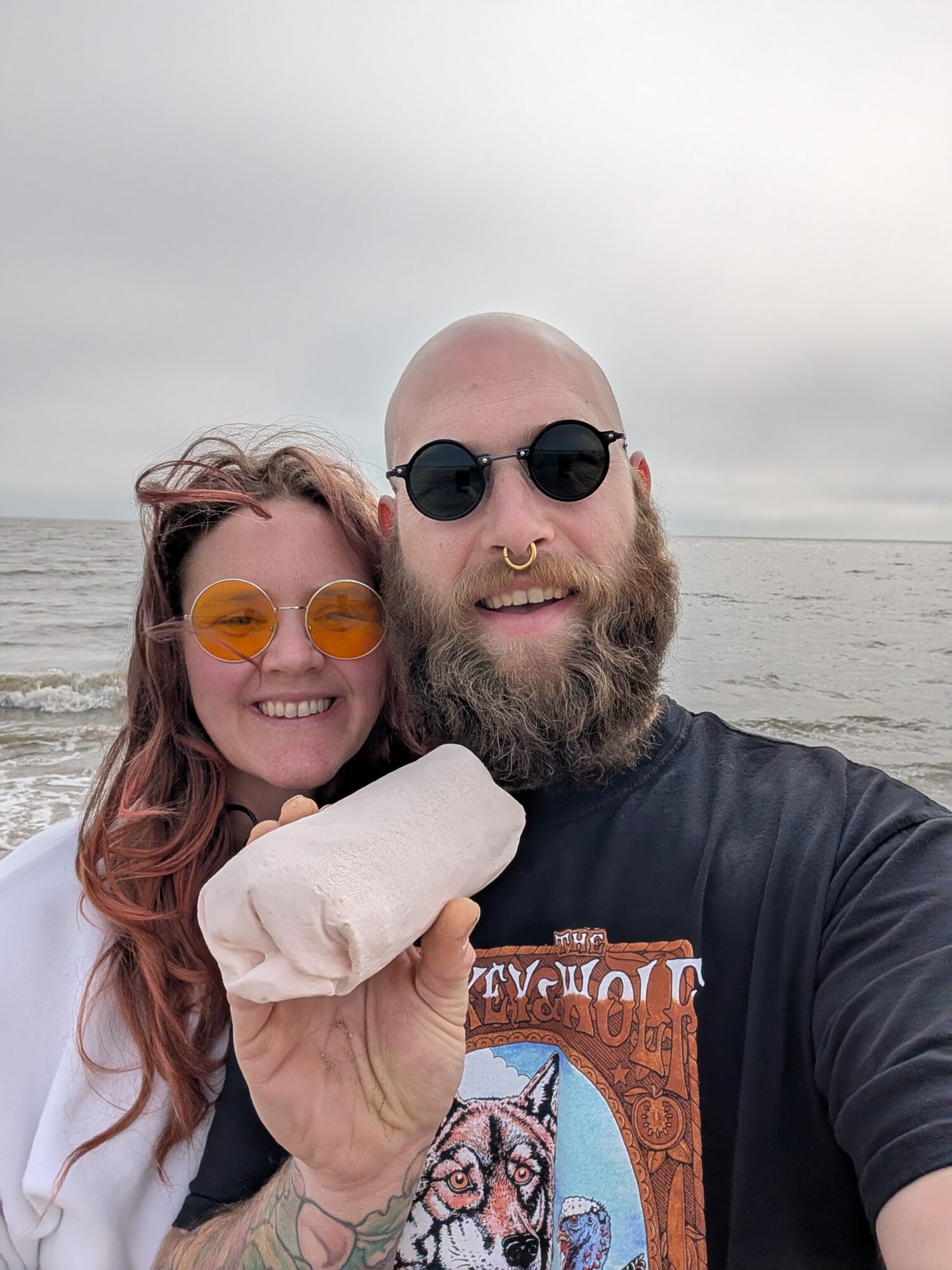 Two people smiling at the camera, wearing sunglasses, holding a large ice cream cone, with a beach and ocean in the background.