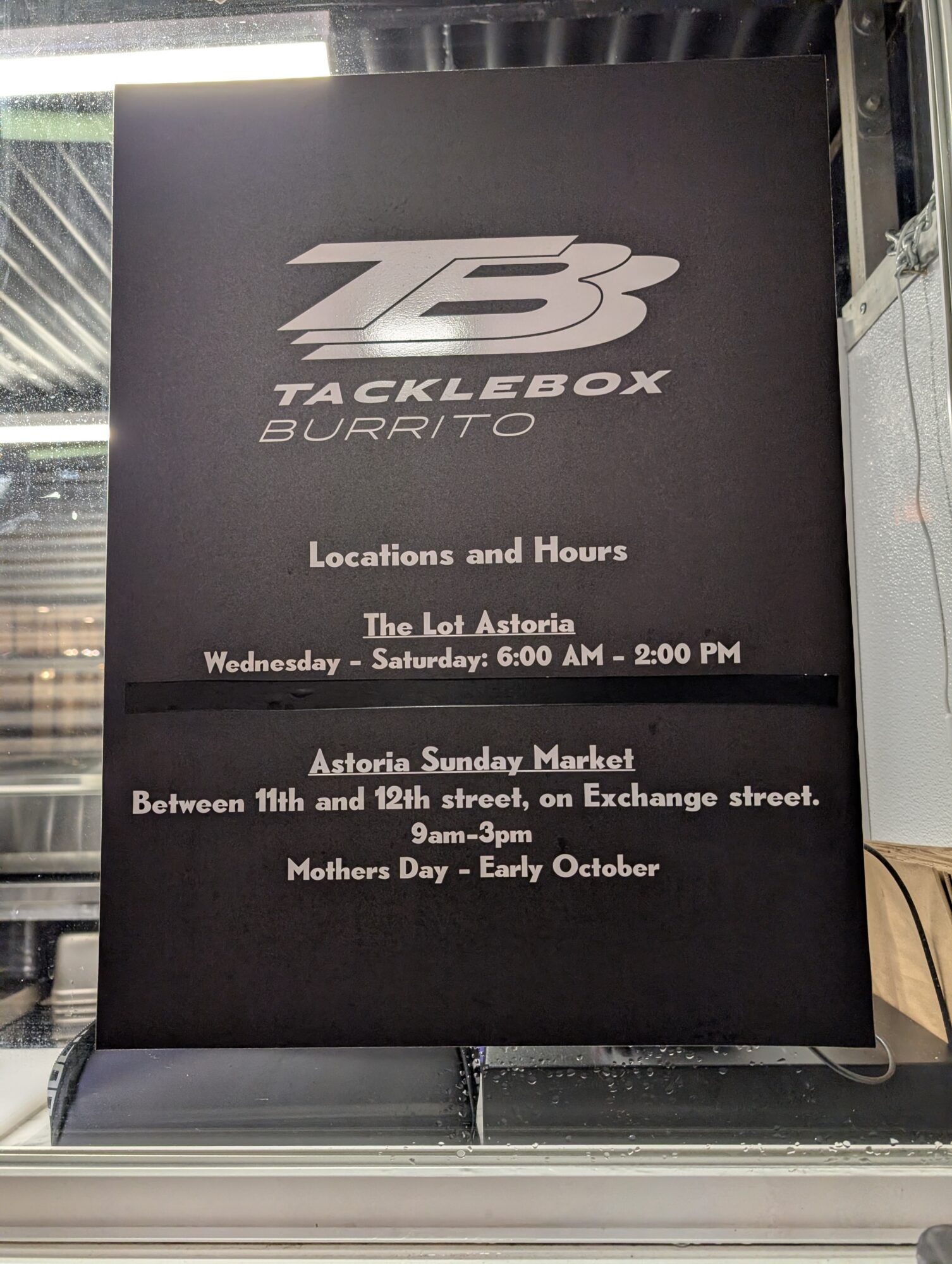 Black sign with white text displaying Tacklebox Burrito's name, location, and hours, including The Loft Astoria and Astoria Sunday Market.