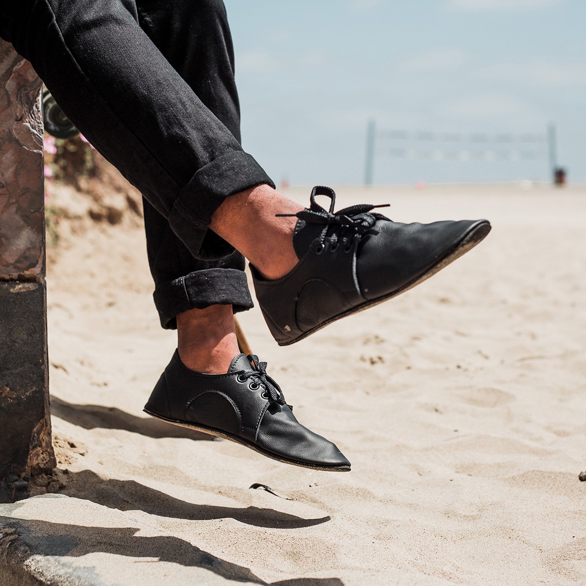 Person sitting on a ledge with legs extended, wearing black shoes and rolled-up black pants, on sandy ground.