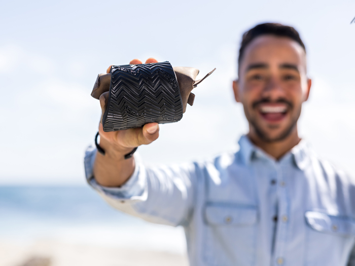 Smiling man holding a small object towards the camera outdoors, with a blurred background of sky and water.