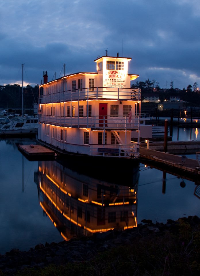 A brightly lit boat docked at a marina during evening with its reflection in calm water, dark cloudy sky overhead.