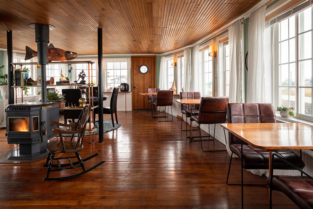Bright room with wooden floors, large windows with curtains, and seating areas with chairs and tables, illuminated by natural light.