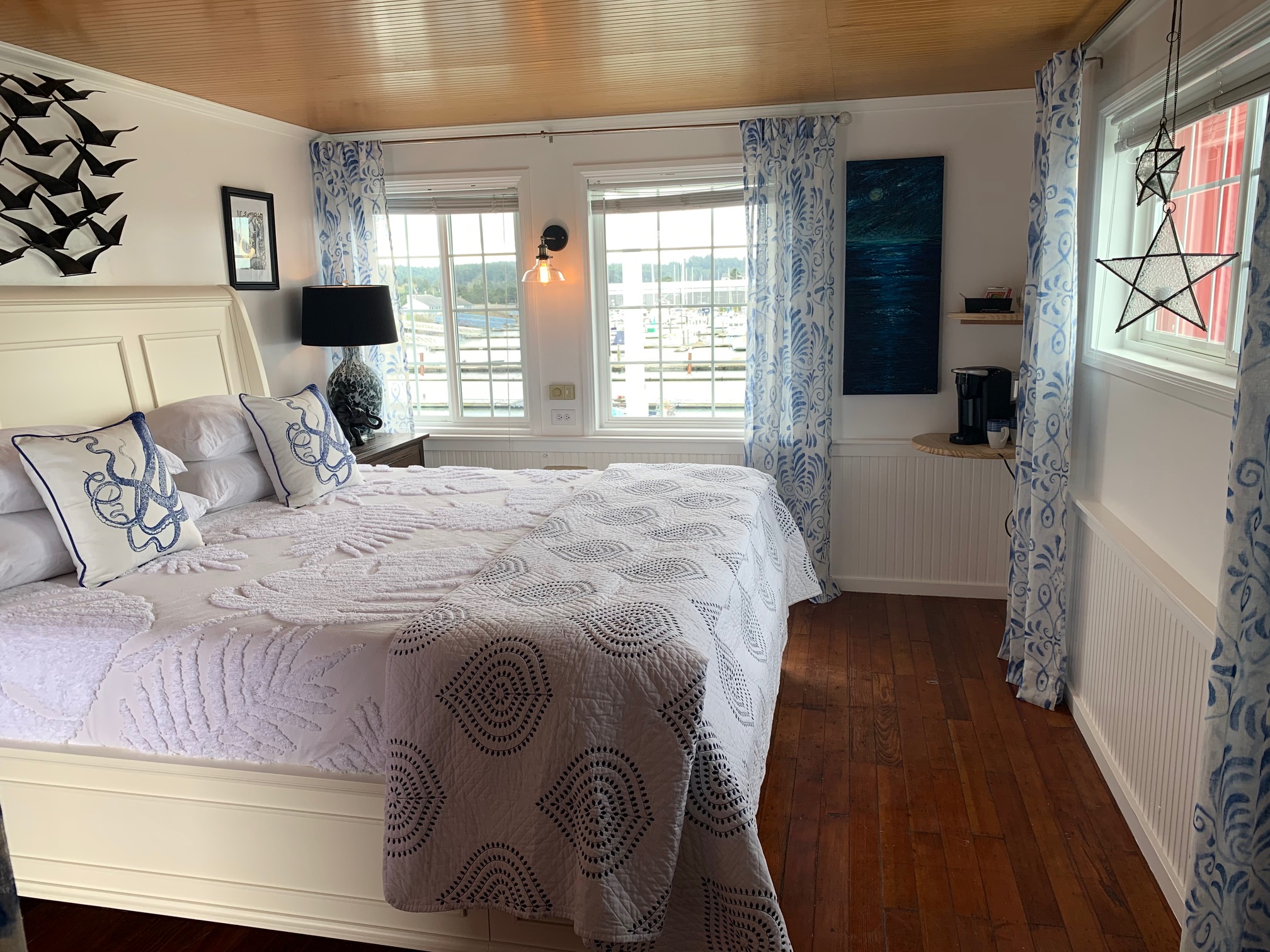 Bedroom with a large bed, white headboard, and pillows, three windows with curtains, wooden ceiling, and hardwood floor.