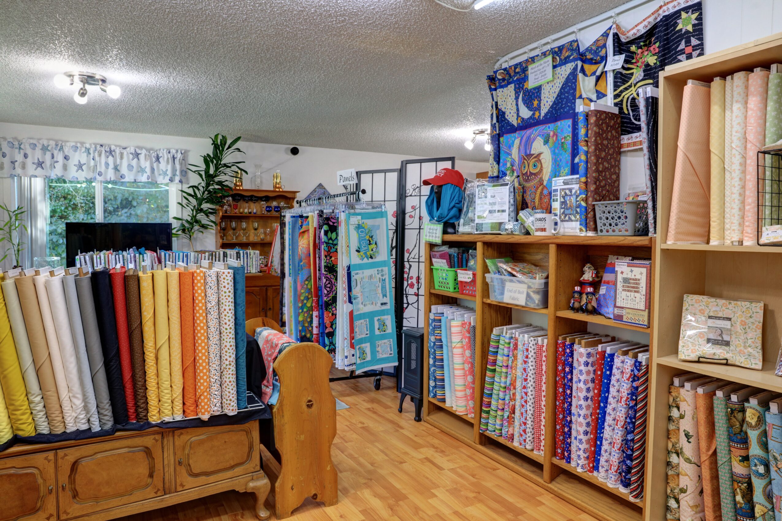 Interior of a fabric store with shelves of colorful fabric rolls, quilting supplies, and a quilt display, wooden flooring, and a window.