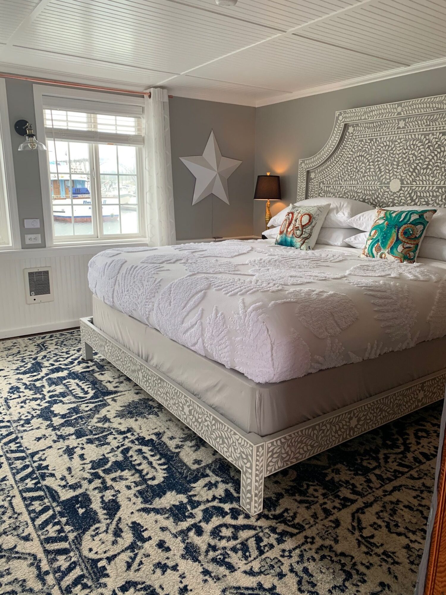 Bedroom with white bed, patterned headboard, window, and blue patterned rug, viewed from the side.