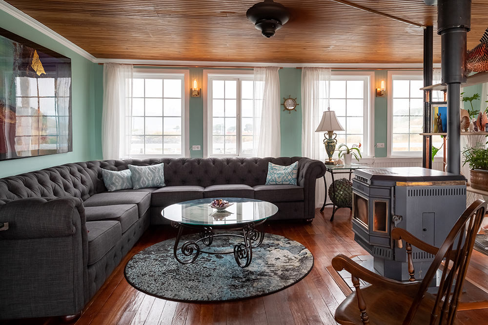 Living room with a large gray sectional sofa, glass coffee table, and wooden floor. Multiple windows with white curtains, wooden ceiling, and various furniture pieces.