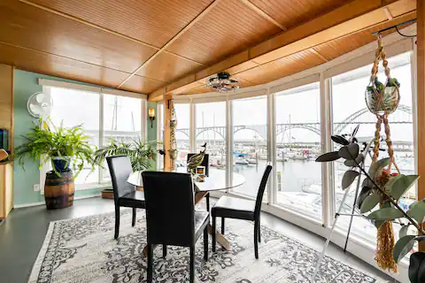 Dining area with four chairs around a table, large windows overlooking a marina, potted plants, and a patterned rug.