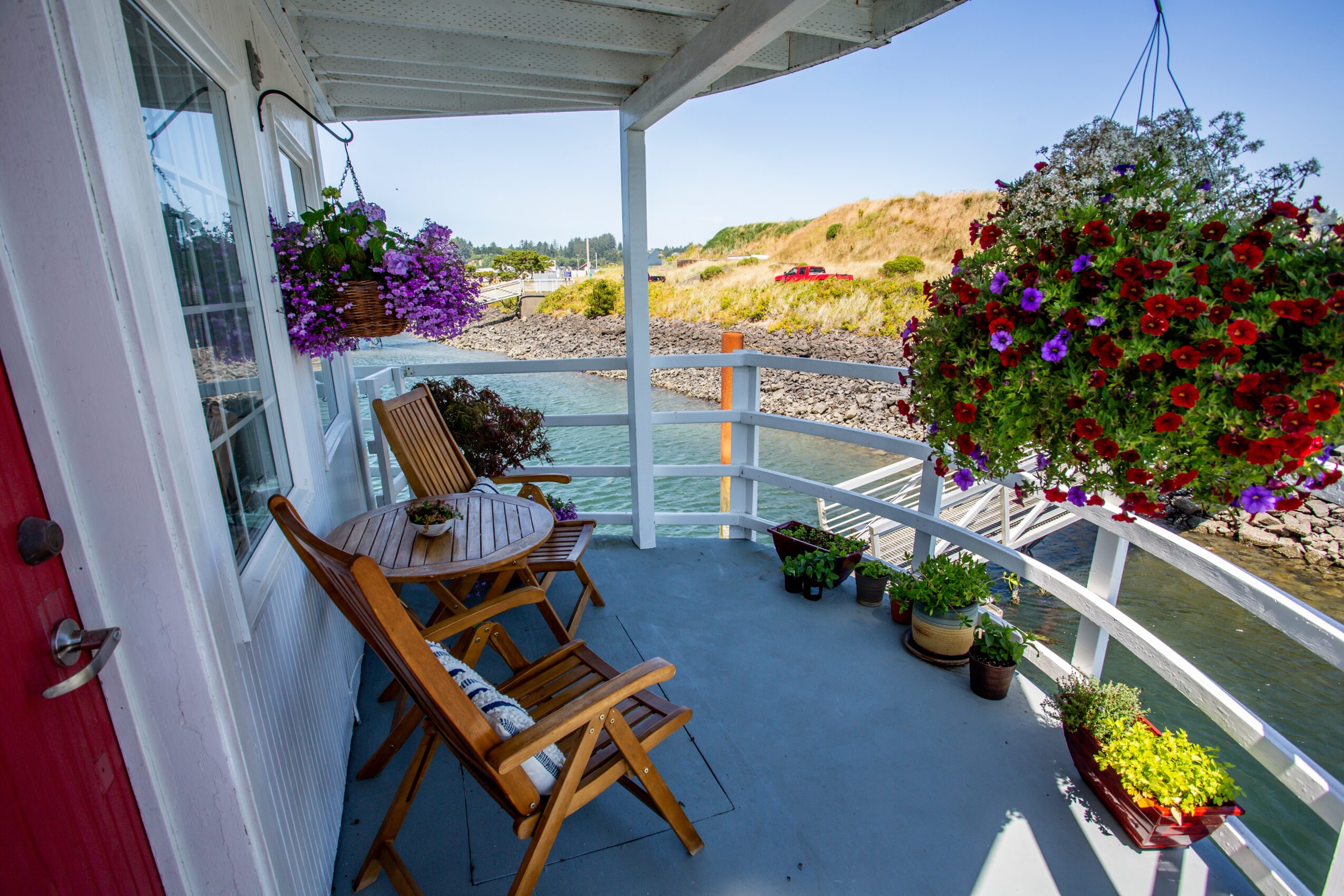 Balcony with wooden chairs, hanging flower baskets, potted plants, overlooking a body of water and landscape.
