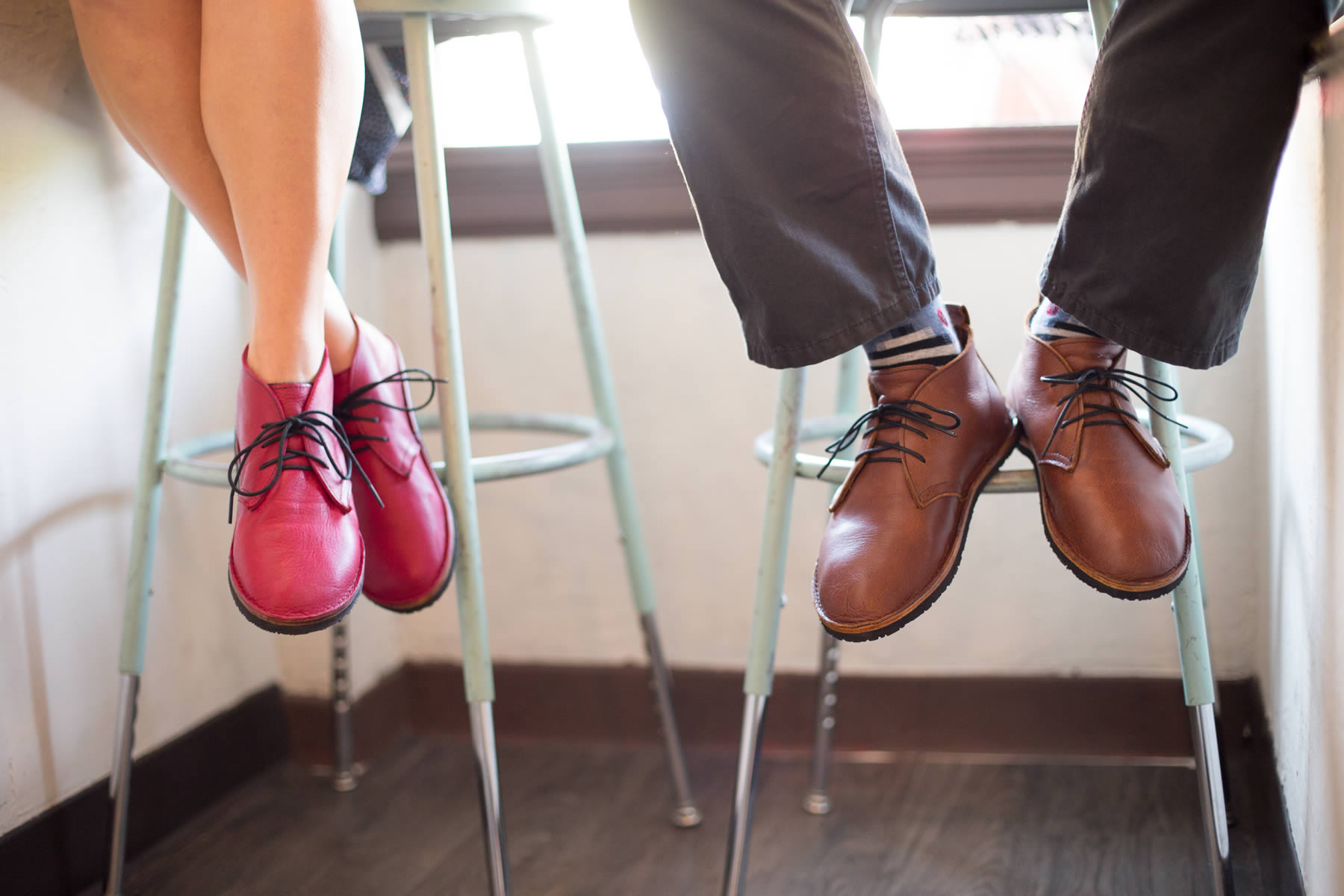 Two people sitting on stools, showing their shoes and legs, with a window in the background.