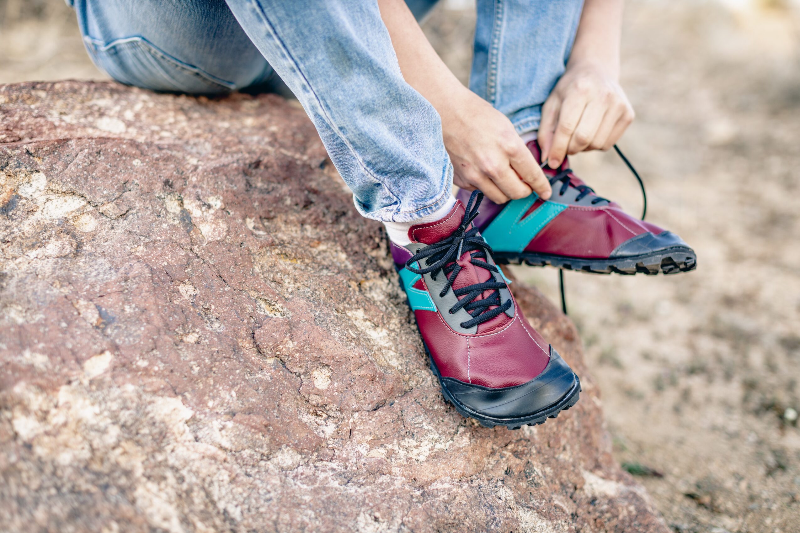 Person tying shoelaces while sitting on a rock outdoors, wearing jeans and colorful hiking shoes.