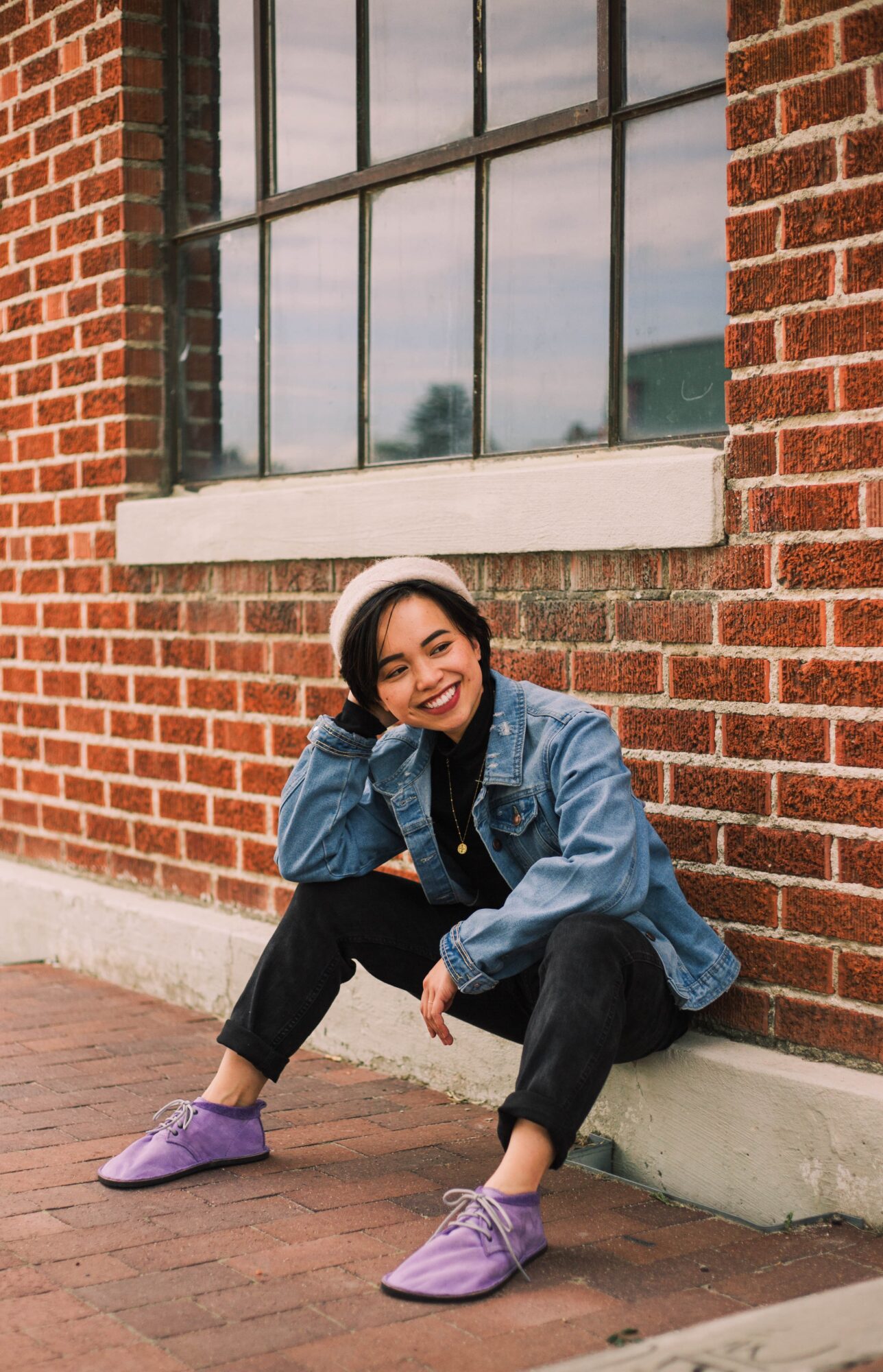 Young woman with headphones smiling, crouching against a brick wall with large window, wearing a denim jacket and purple shoes.