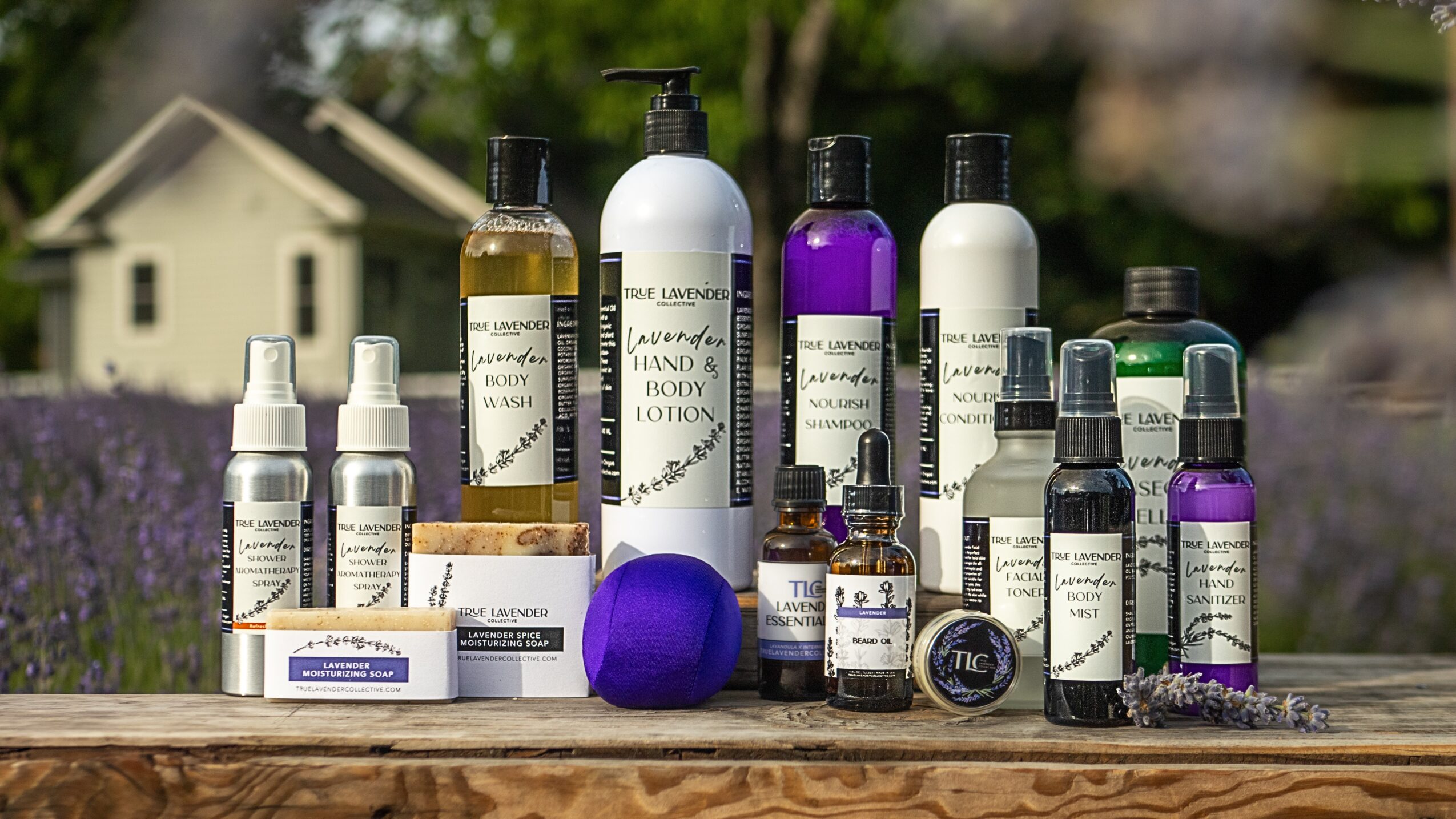 Collection of skincare and body products with bottles, jars, and boxes outdoors on a wooden surface, with a house and garden in background.