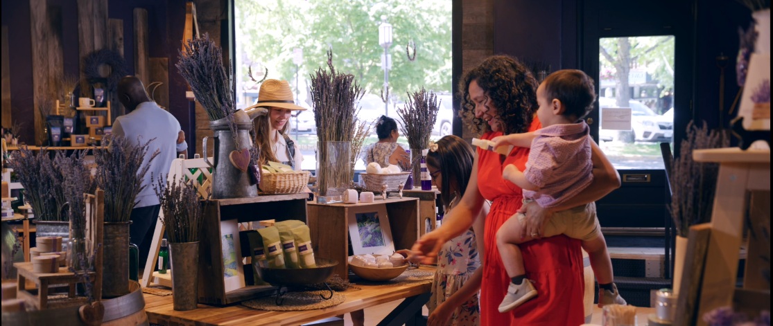 People shopping in a store with wooden shelves and large windows, a woman holding a child, and others browsing displays.