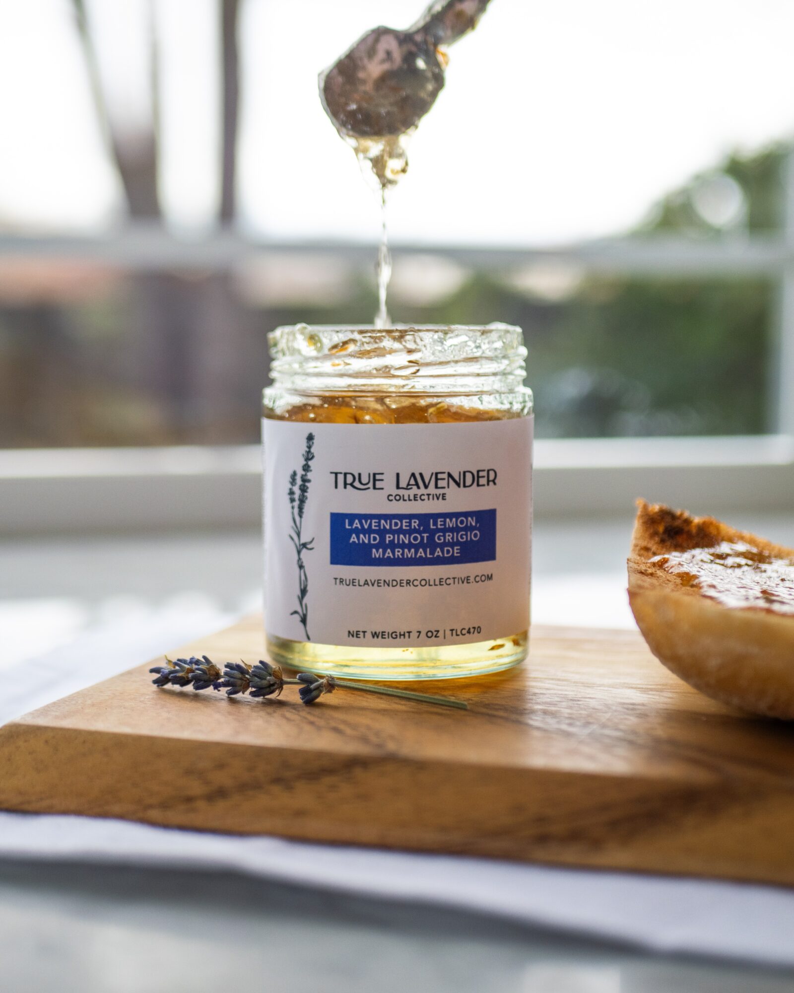 Open jar of lavender honey with honey dripping from a spoon above it, on a wooden surface with a pie nearby.