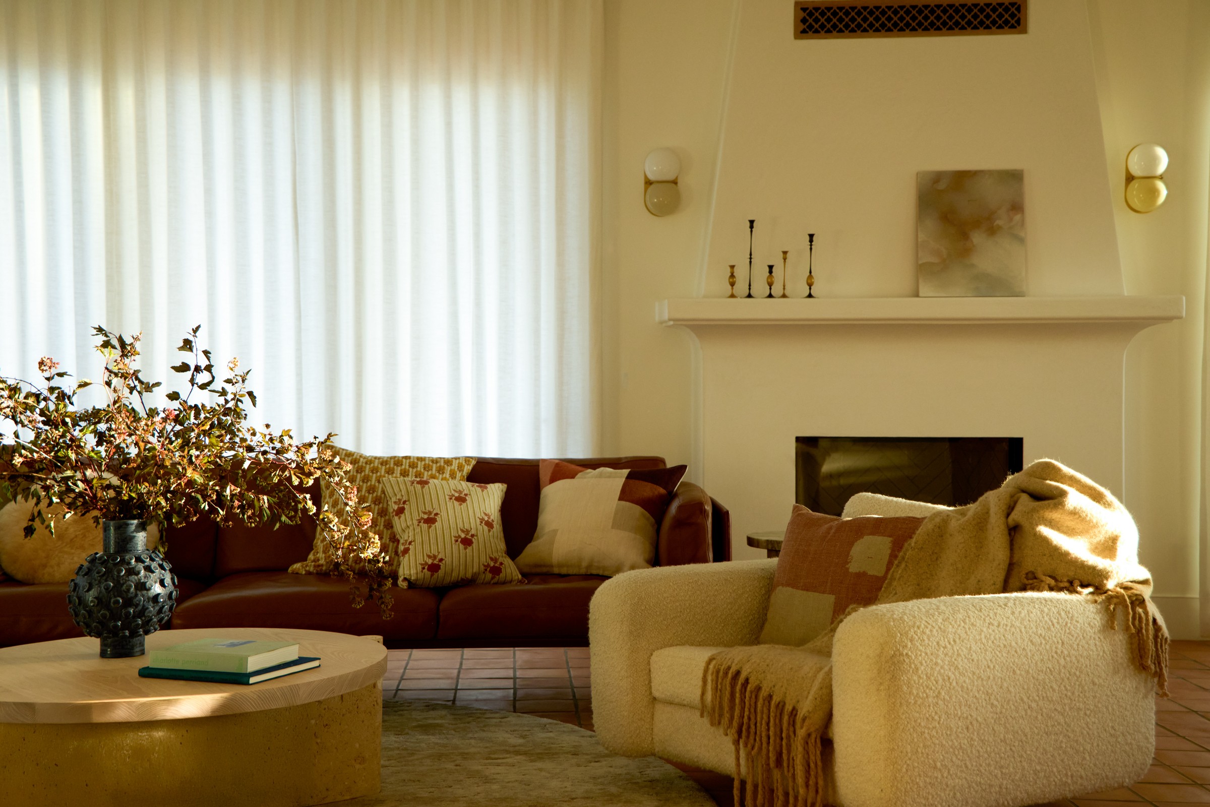 Living room with a sofa, armchair, fireplace, and window with curtains, decorated with cushions and flowers.