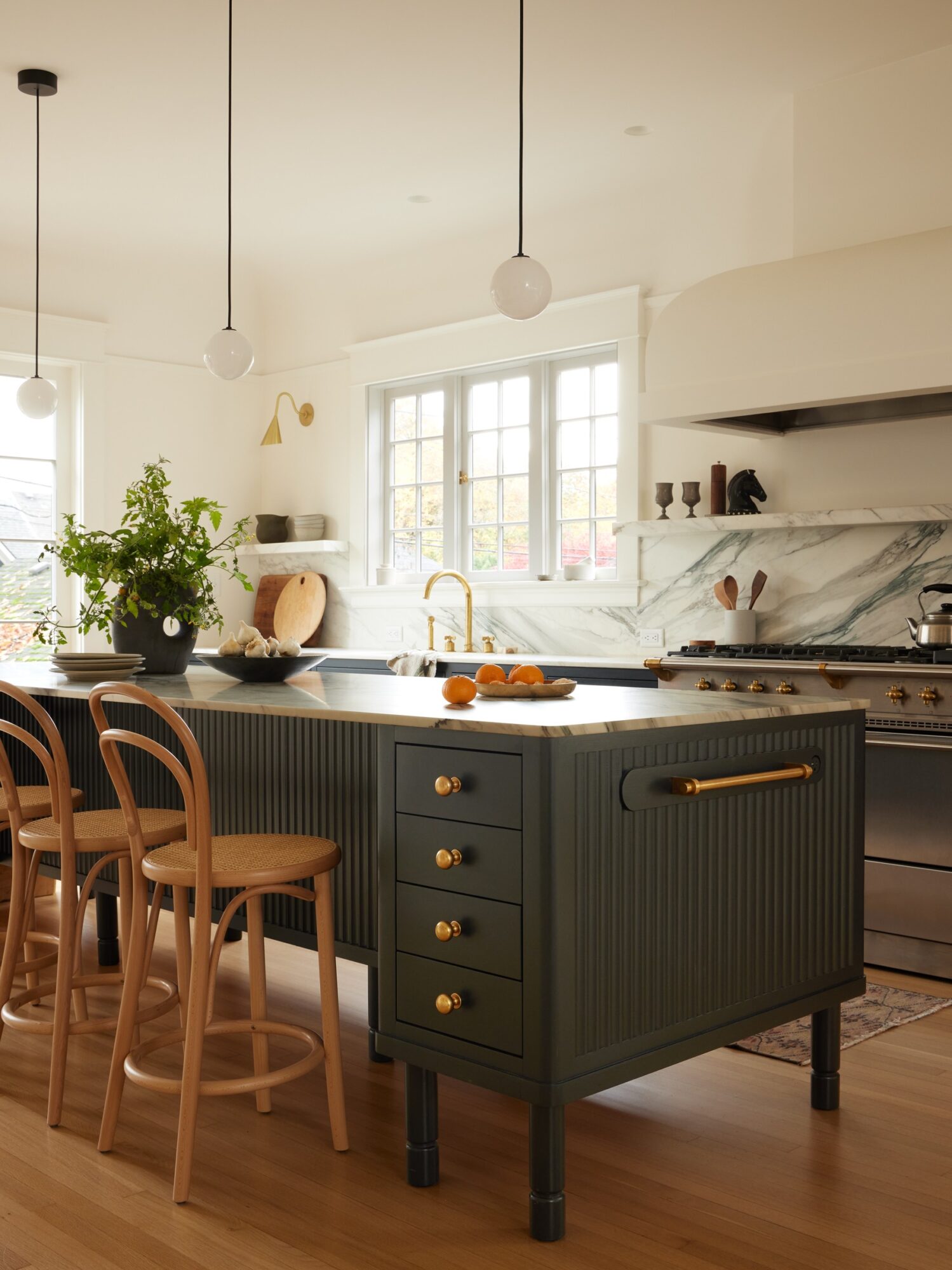 Kitchen with dark island, wooden chairs, white cabinets, and large window, bright and airy space with plants and decor.