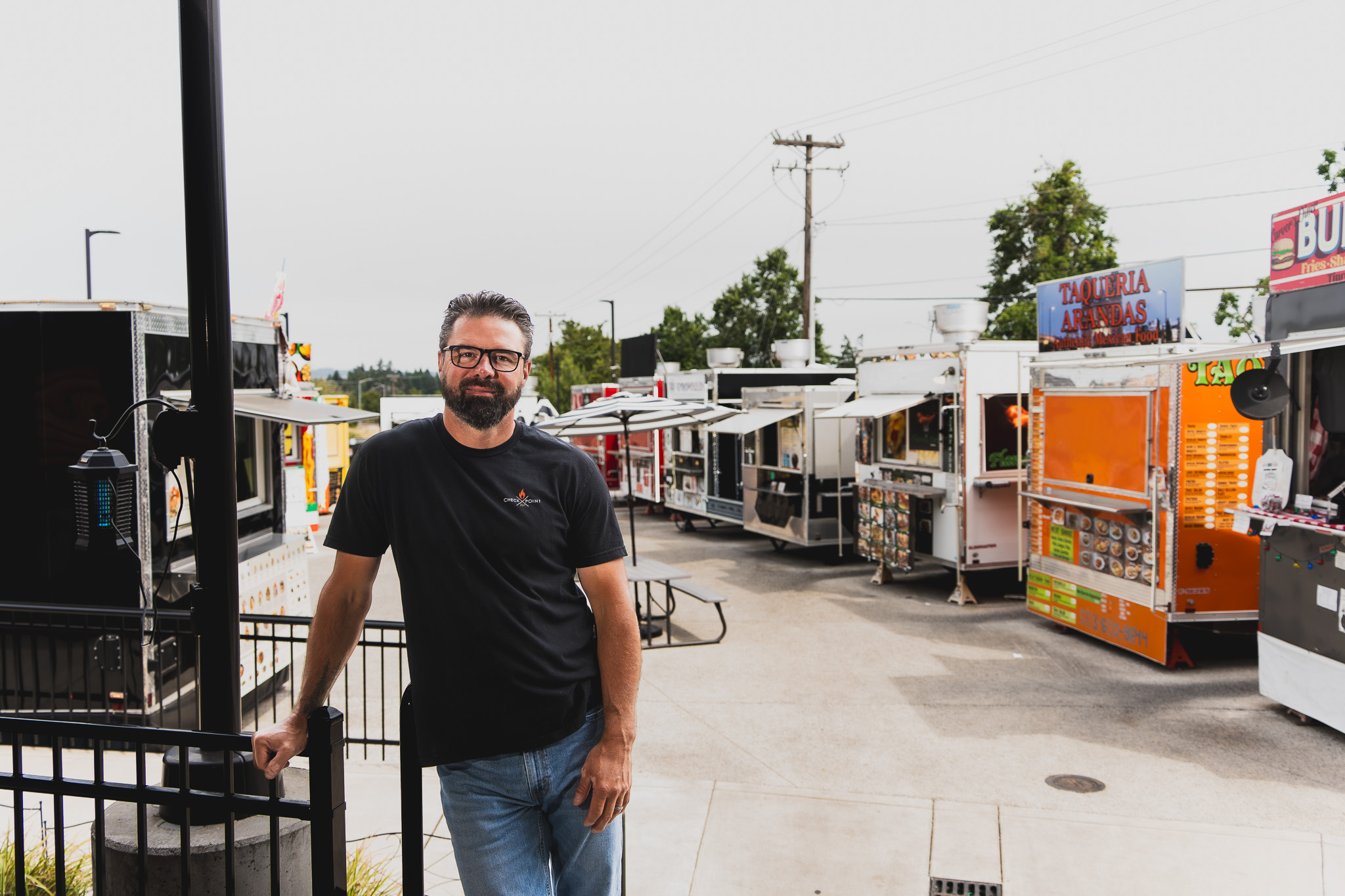 Man with glasses and beard standing outdoors near food trucks, smiling, wearing a black shirt and jeans.