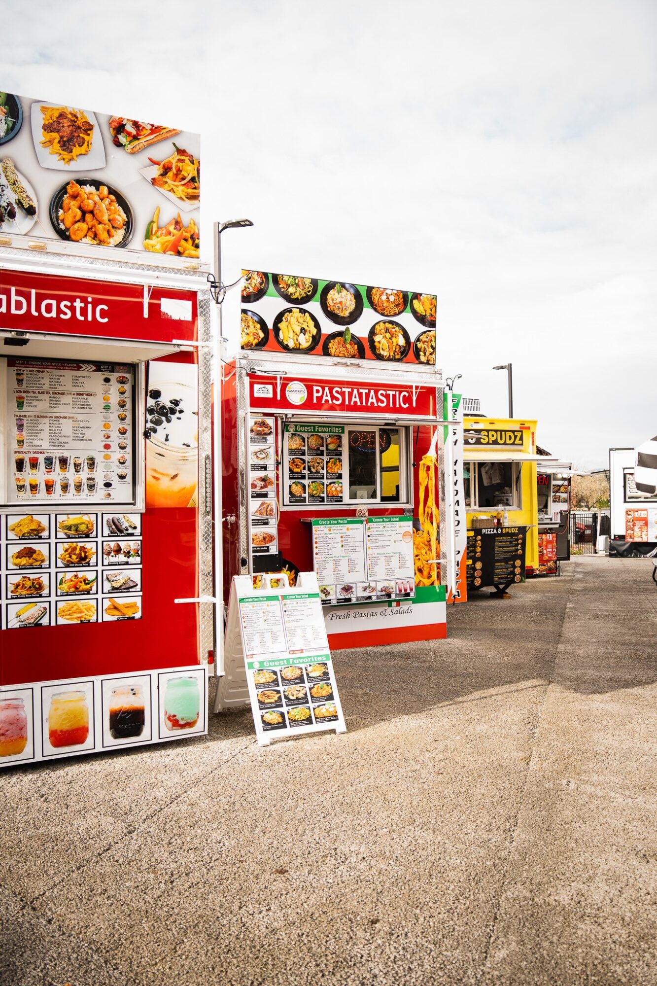 Food vending stalls with colorful menus and food images, set outdoors on a paved area under a cloudy sky.