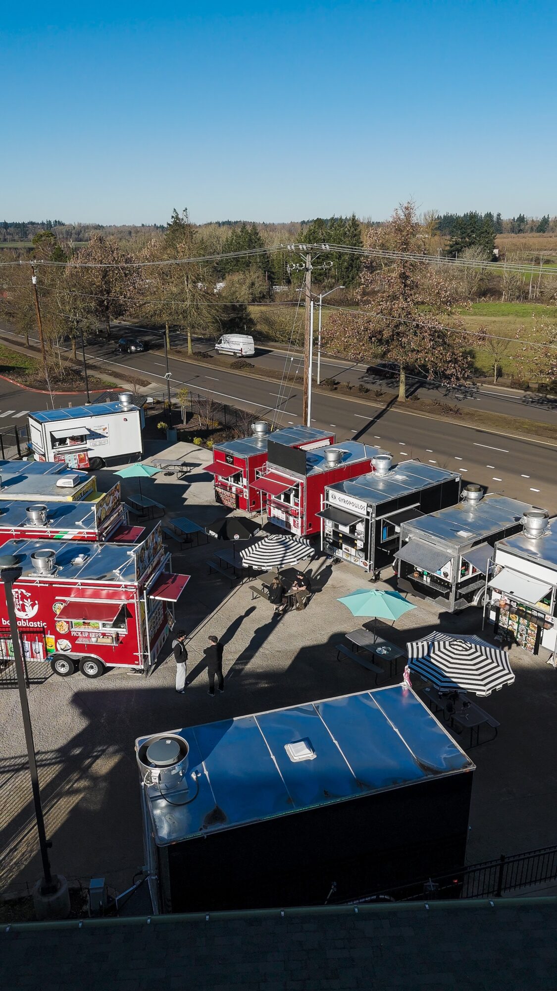 Food trucks parked in a lot with trees and a road in the background, sunny day, shadows cast on the ground.