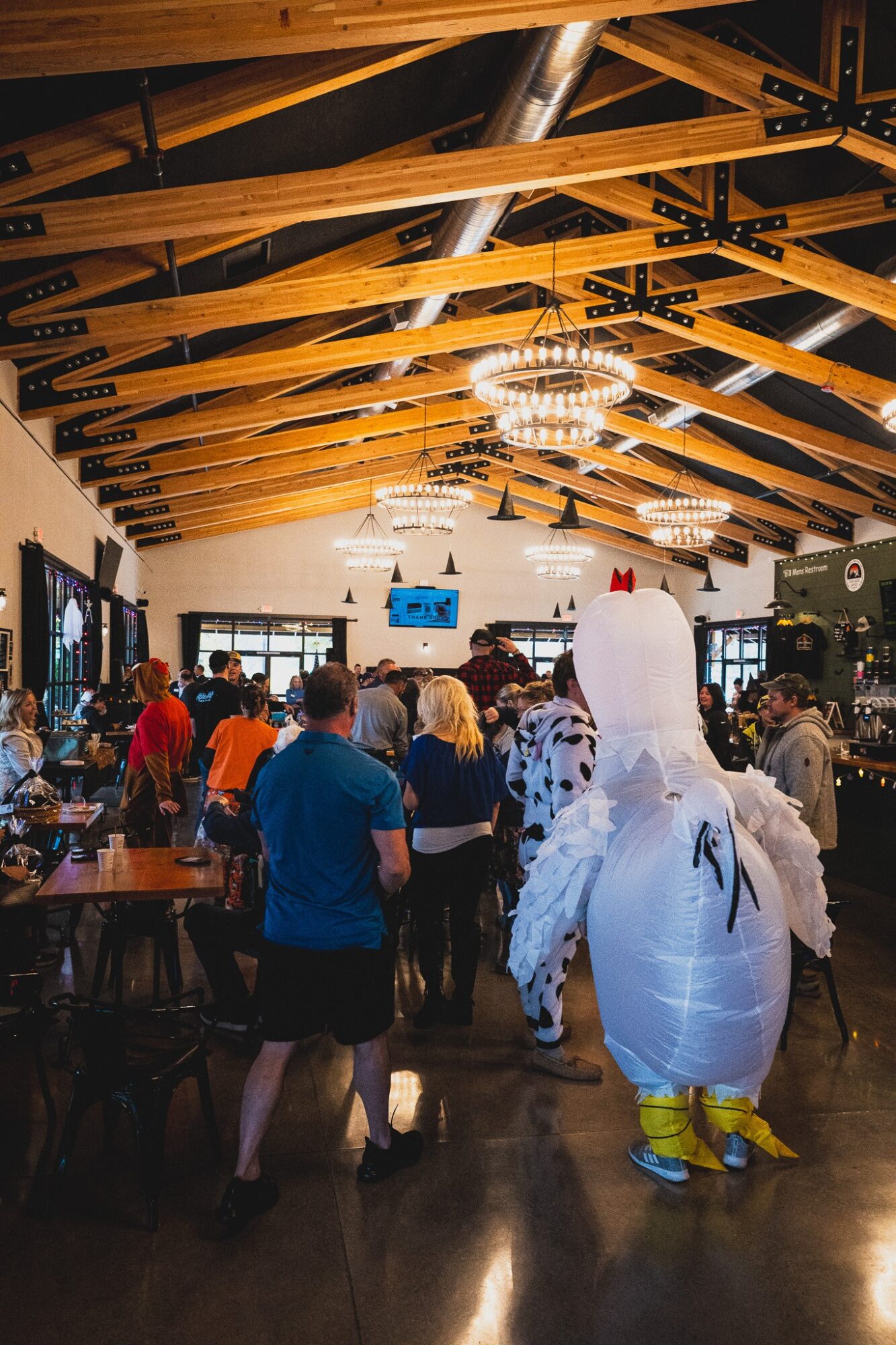 People gather in a decorated indoor space with wooden beams, chandeliers, and a person in a bird costume, celebrating.
