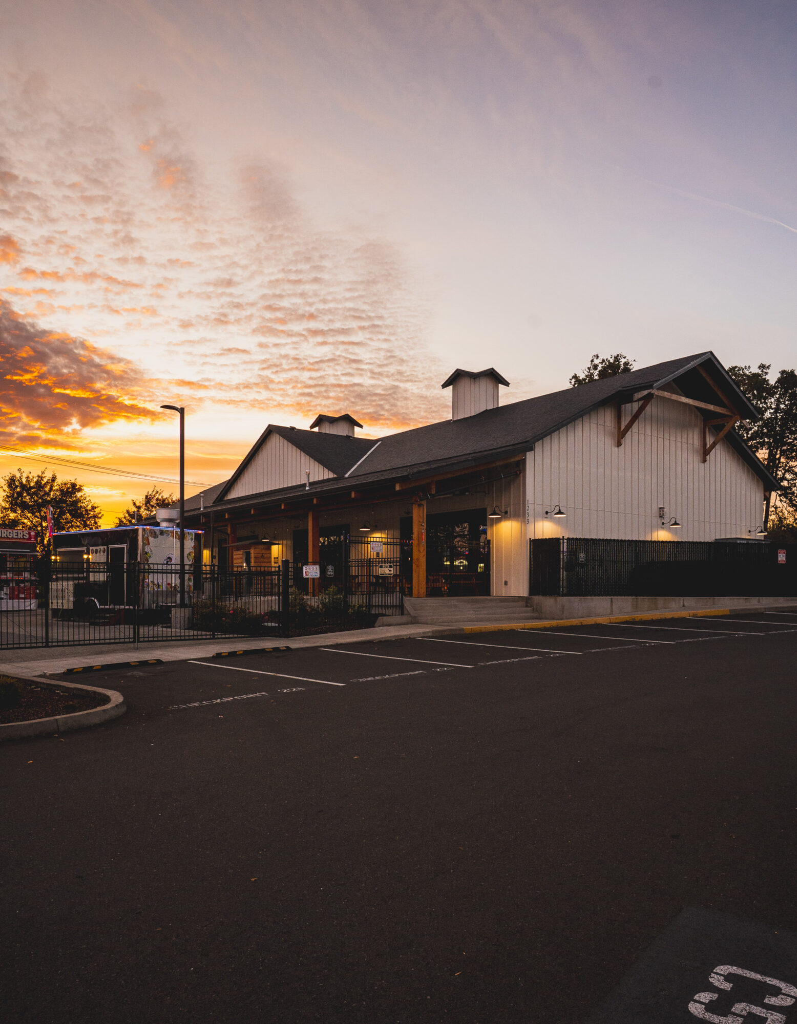 Building with a sloped roof and chimney, sunset sky with clouds, street in foreground, trees in background.