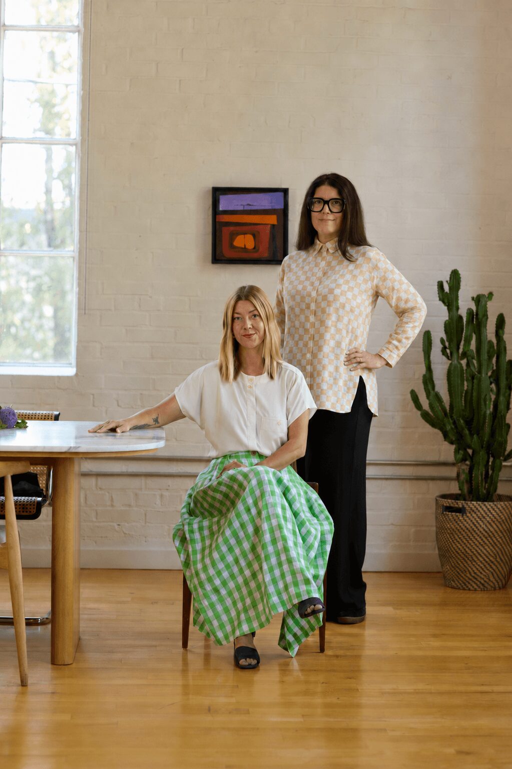 Two women in a room with a large window, one seated at a desk and the other standing beside her, with a cactus plant nearby.