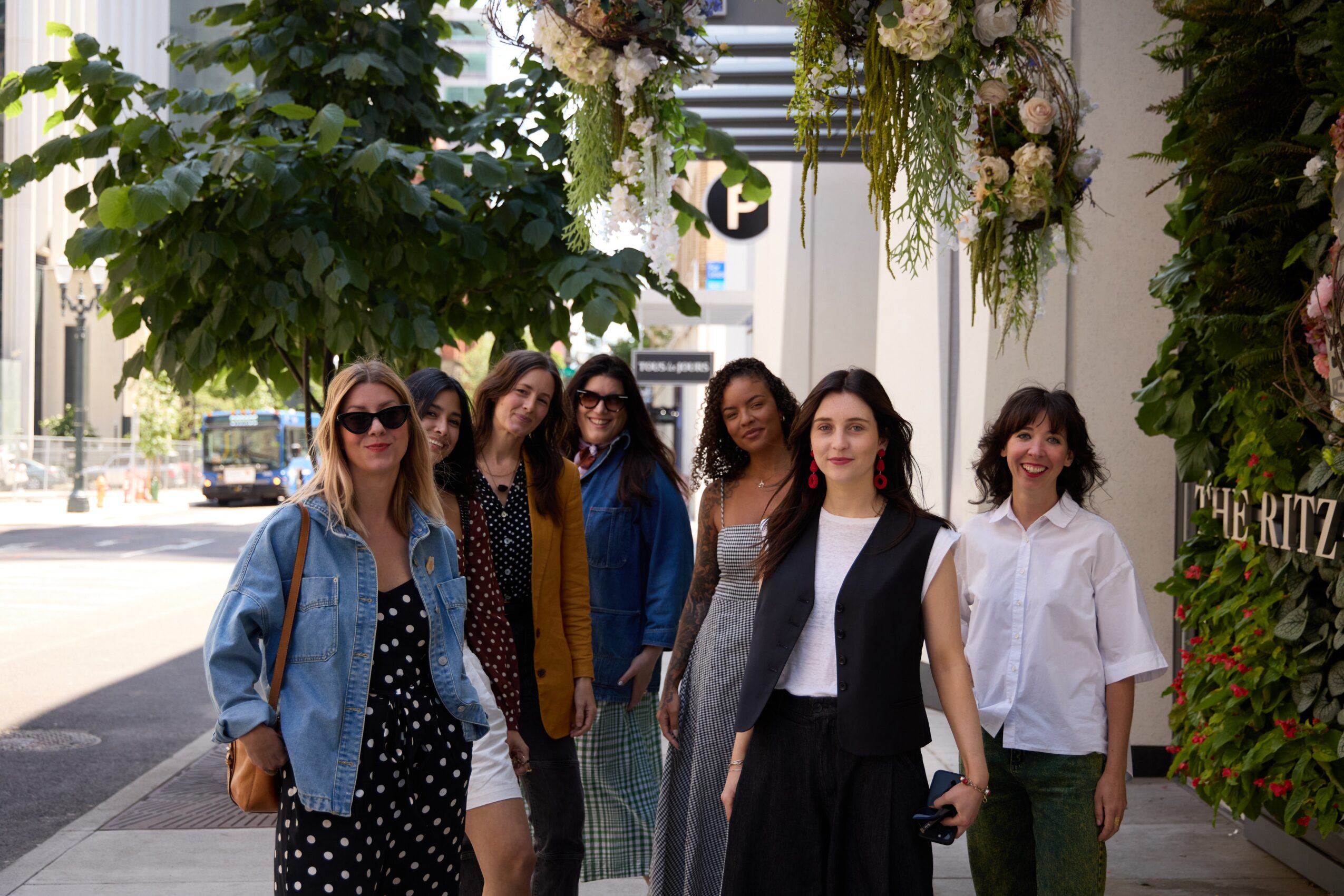 Group of six women standing outdoors under hanging floral decorations, smiling, with trees and buildings in the background.