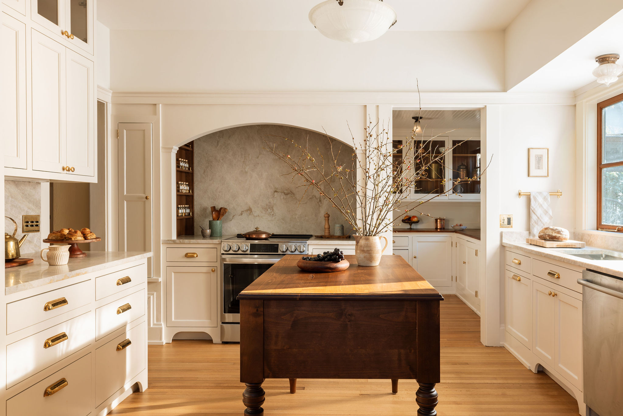 Kitchen with white cabinets, wooden floor, central wooden table, and window with natural light.