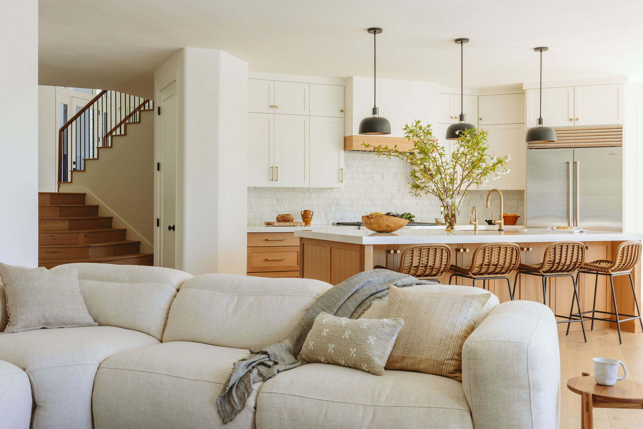 Open-concept kitchen and living room with white cabinets, wooden accents, and a large island with bar stools, and a staircase in the background.