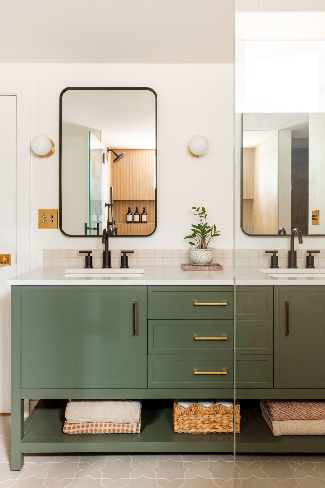 Double bathroom vanity with green cabinets, two mirrors, and a potted plant between sinks, with a basket and towel below.