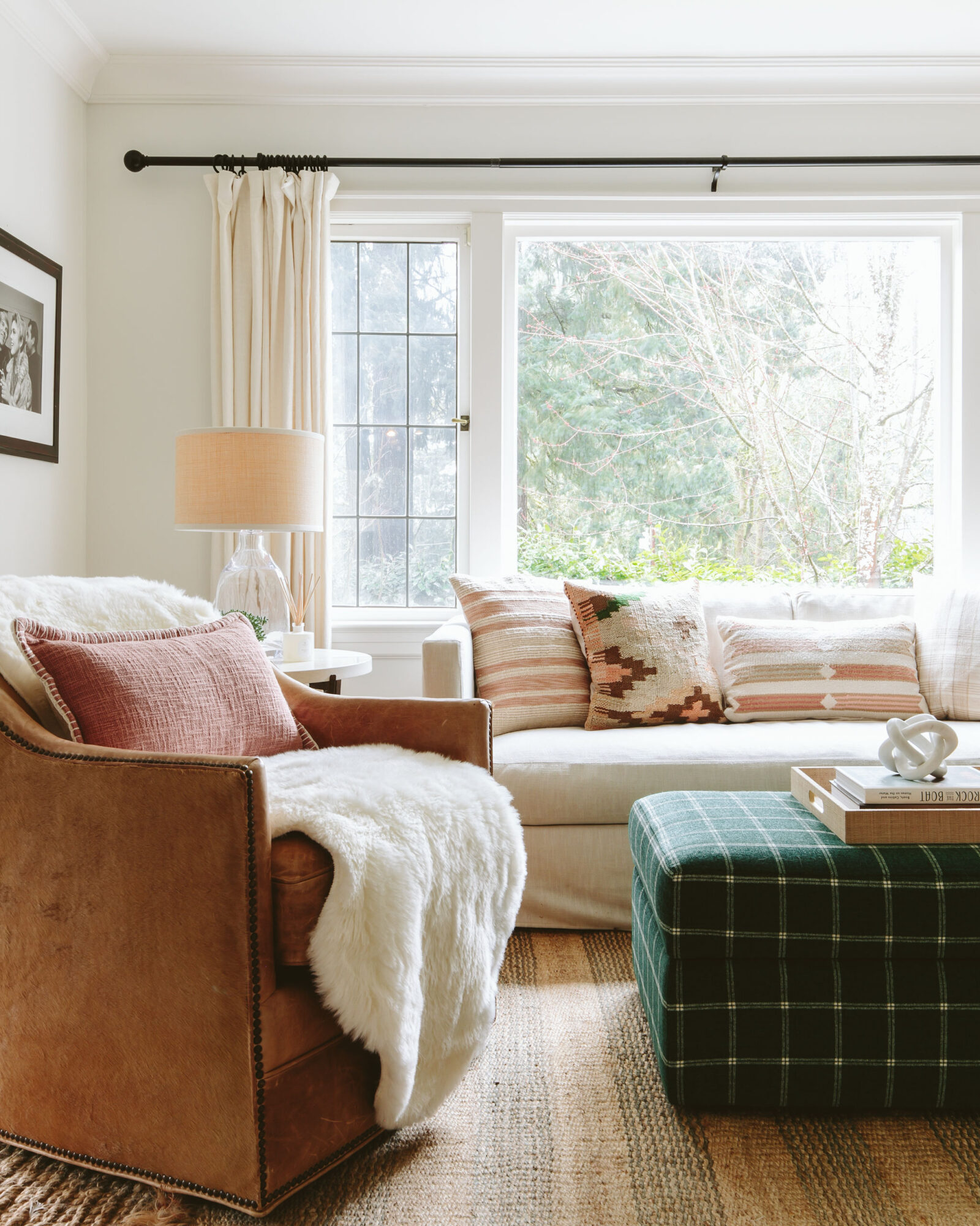 Living room with a window, sofa with pillows, armchair with a throw, and a plaid ottoman, natural light, neutral colors.