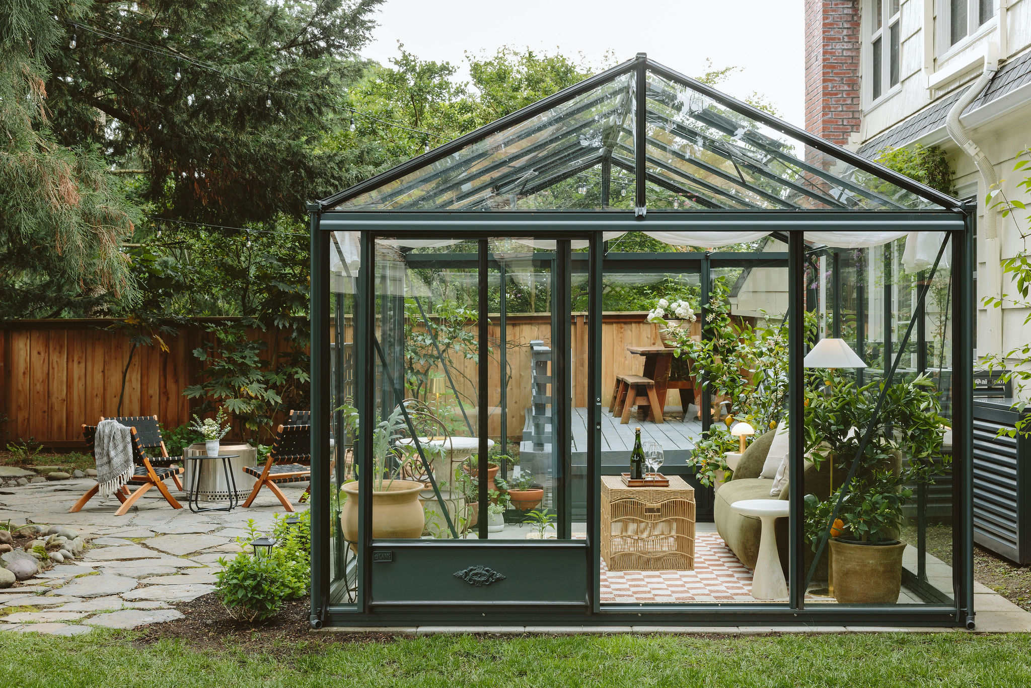 Glass greenhouse with plants and furniture inside, outdoor garden setting with trees and a wooden fence.