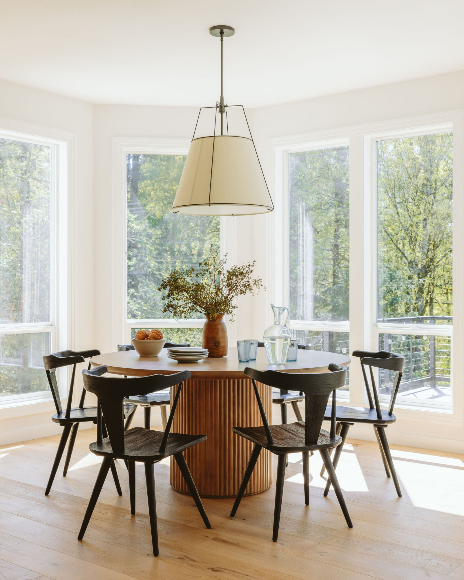 Round dining table with four chairs in a bright room with large windows and a hanging light fixture above.