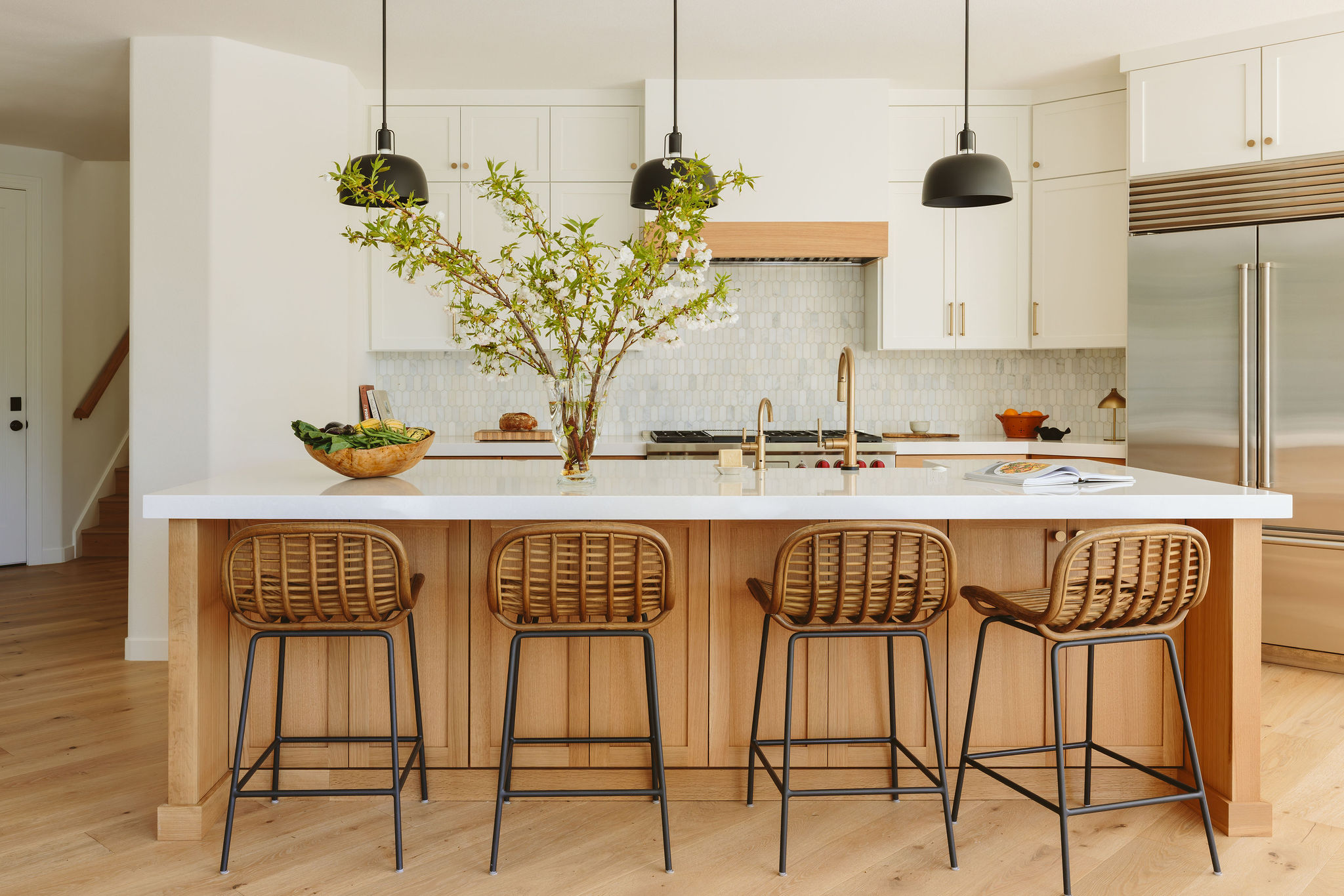Kitchen with a large island, four wicker bar stools, a tree in a bowl, and modern white cabinets.