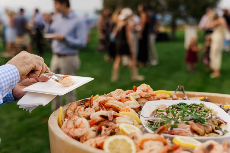 Person holding a plate of food at an outdoor event with a large seafood platter in foreground and people in background.