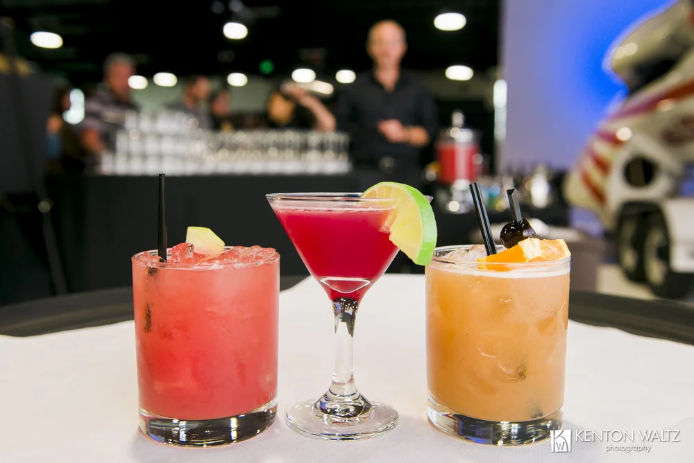 Three colorful cocktails on a bar counter with blurred background of bar scene and people, garnished with fruit slices.