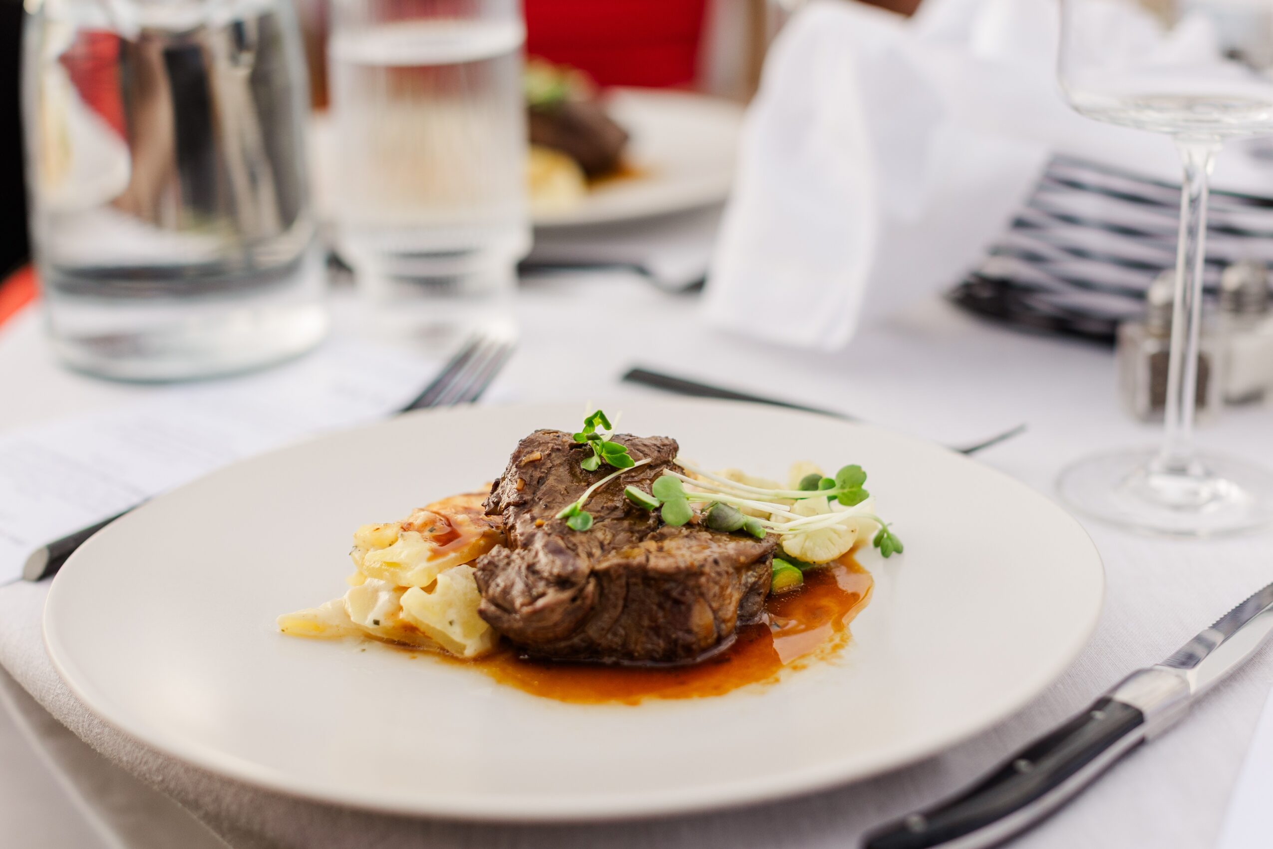 Plate with cooked meat, mashed potatoes, and green garnish on a table with glasses and utensils