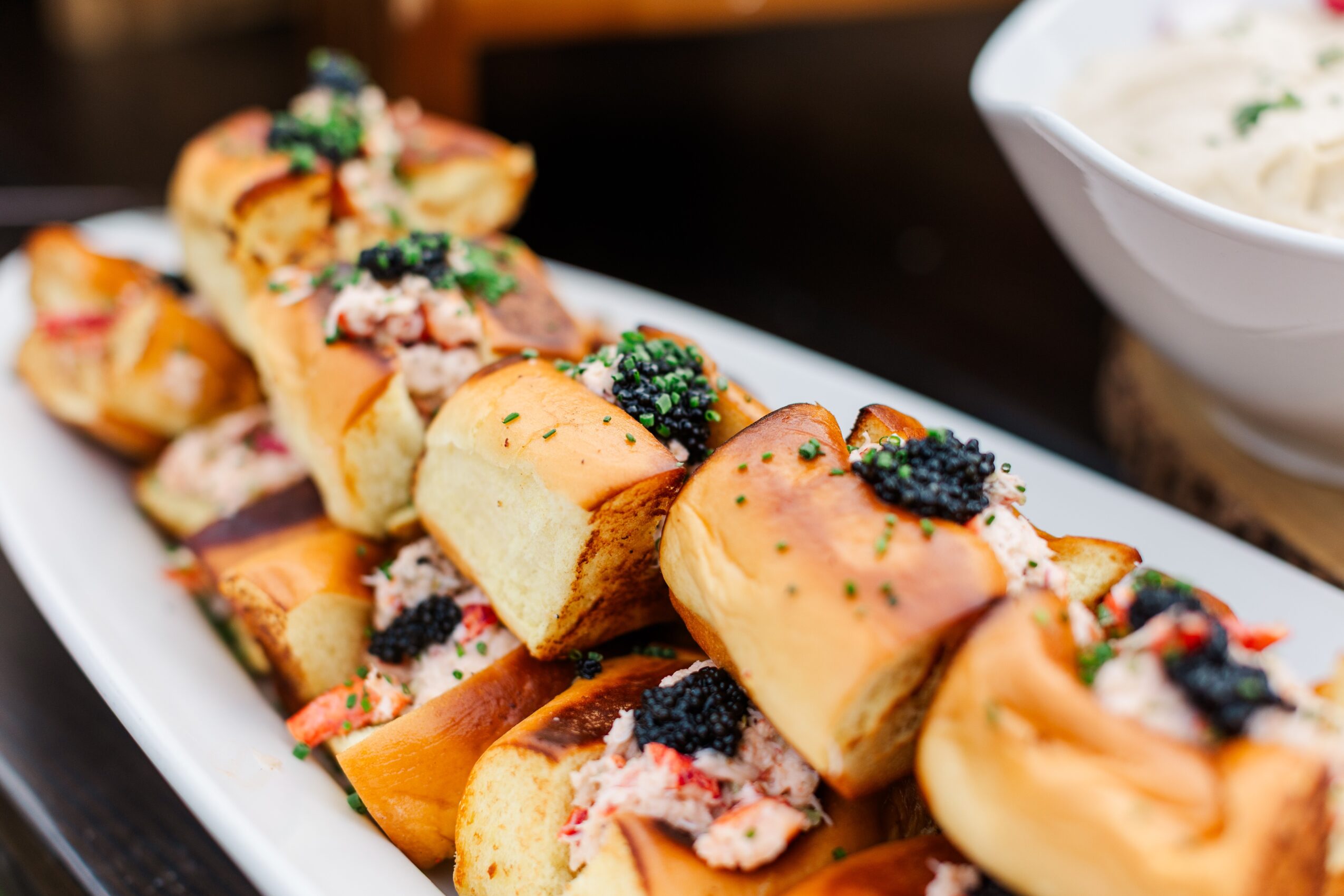Slices of bread topped with caviar, garnished with herbs, arranged on a white platter, with a bowl of soup in the background.