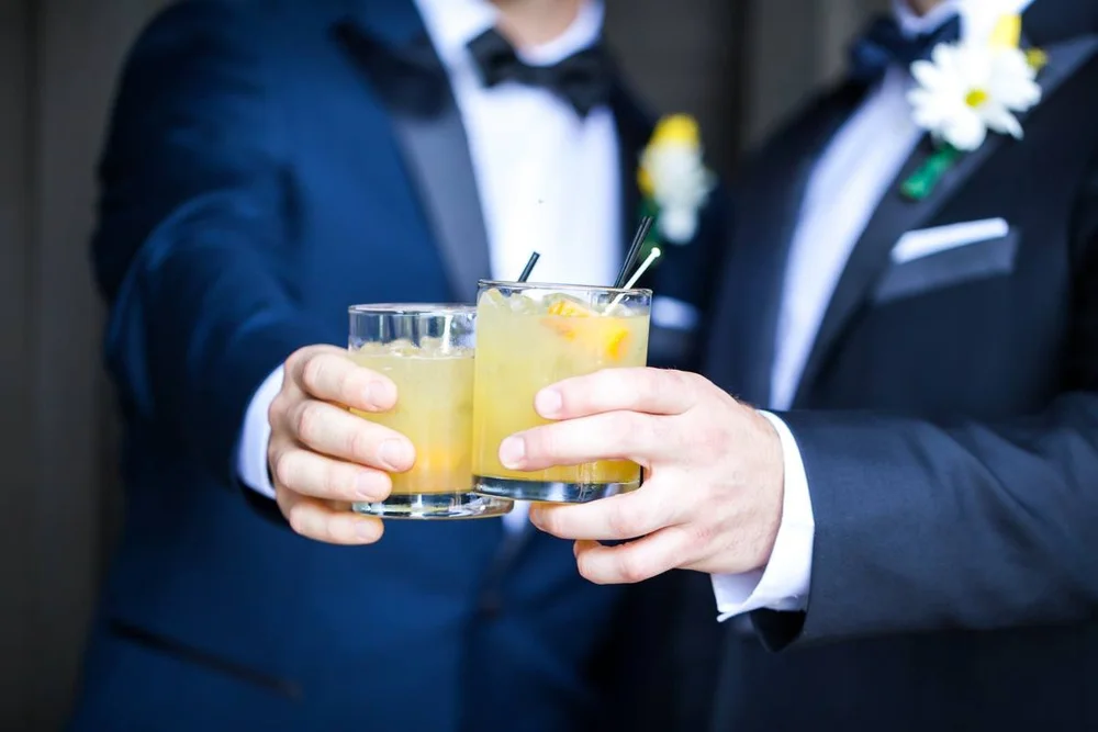 Two people in tuxedos holding glasses of yellow cocktail with lemon slices, celebrating together.