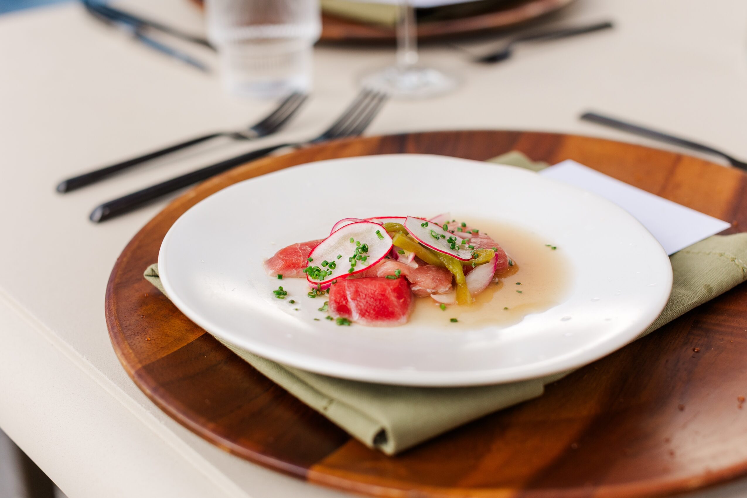 Plate with sliced radishes, herbs, and sauce on a white dish, placed on a wooden tray with a napkin underneath.