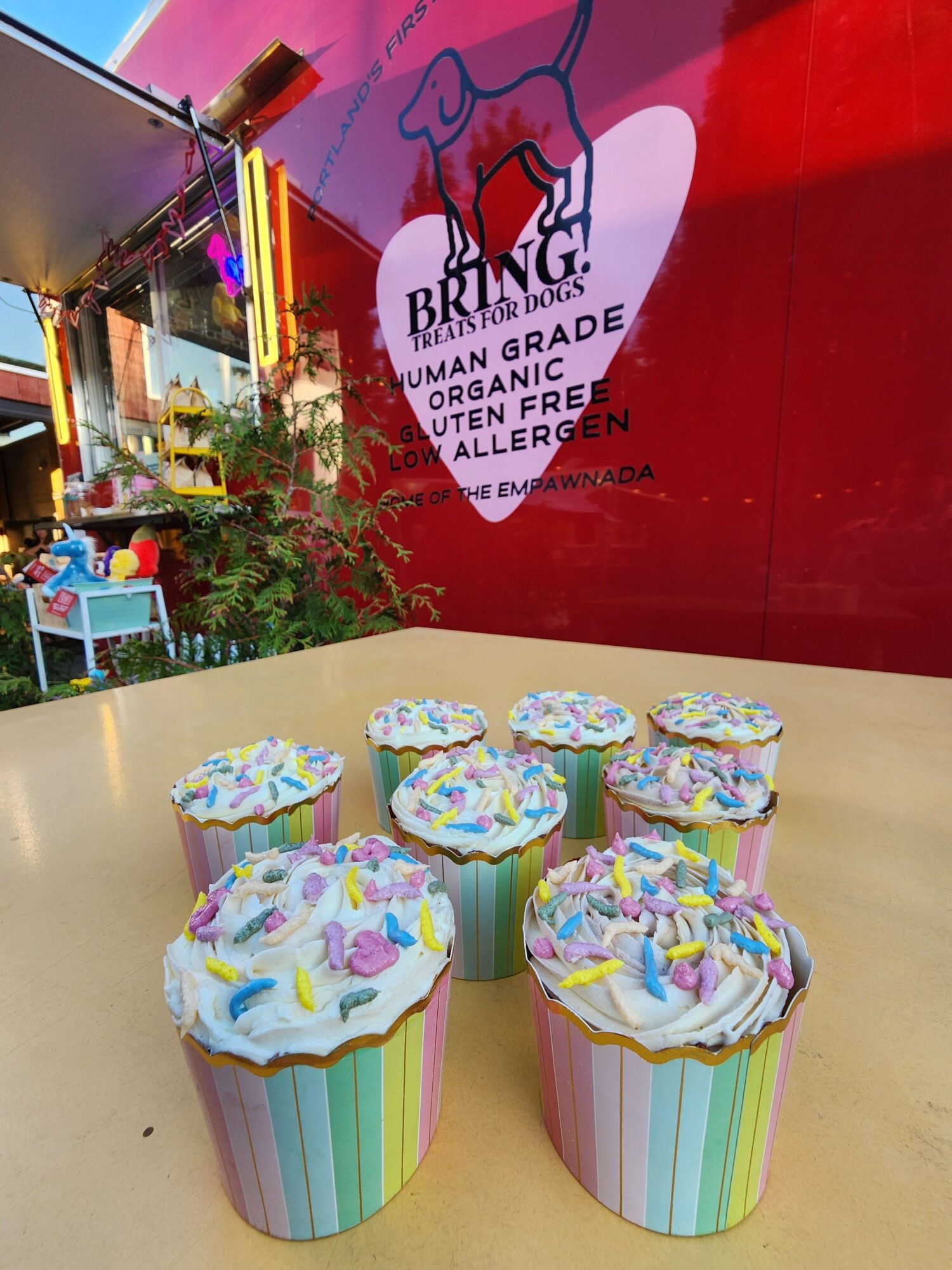 Cupcakes with colorful frosting and sprinkles on a table, with a red wall and a sign in the background.