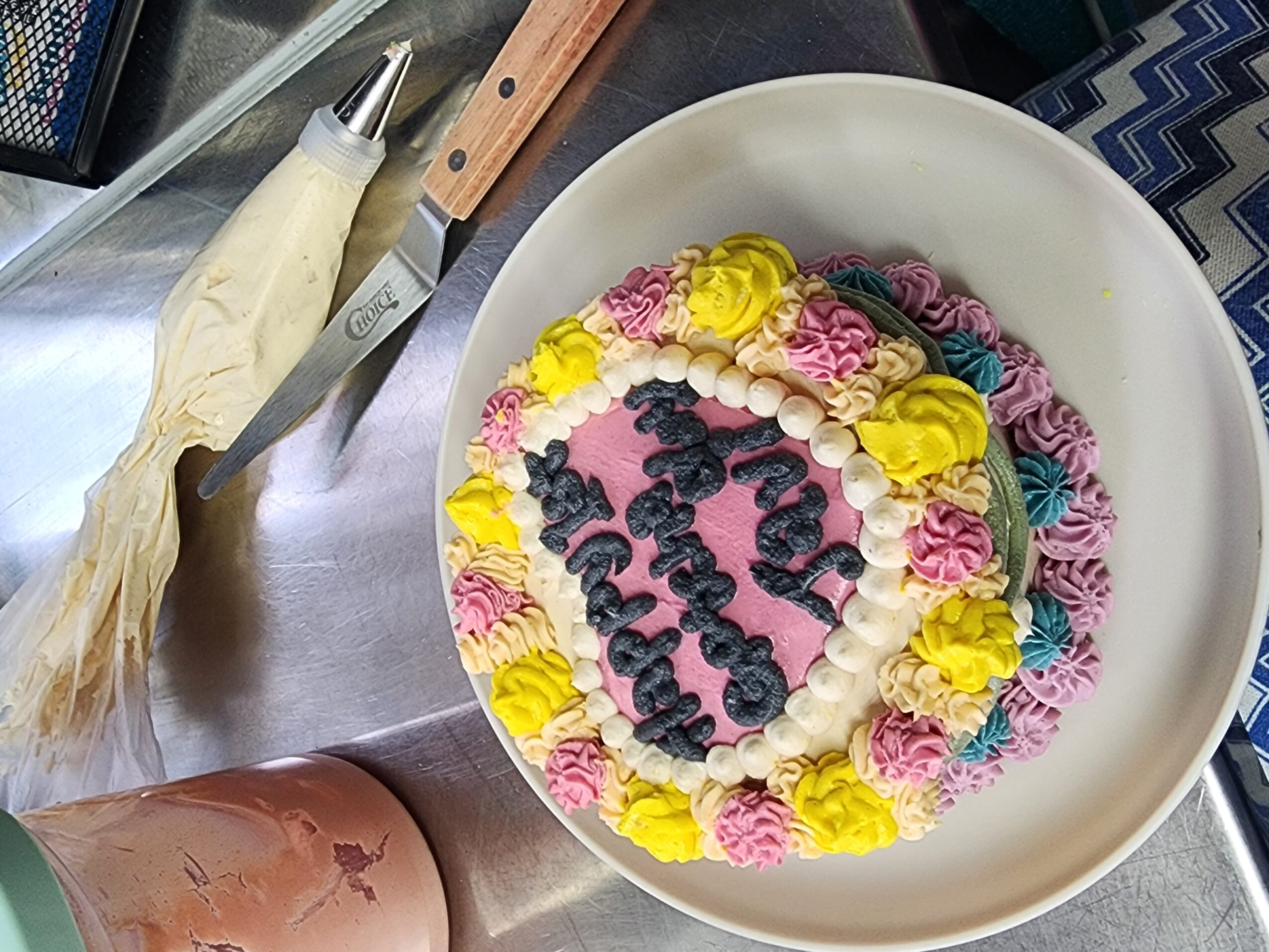 Round cake with colorful frosting flowers and writing in the center, on a white plate, with kitchen utensils nearby.