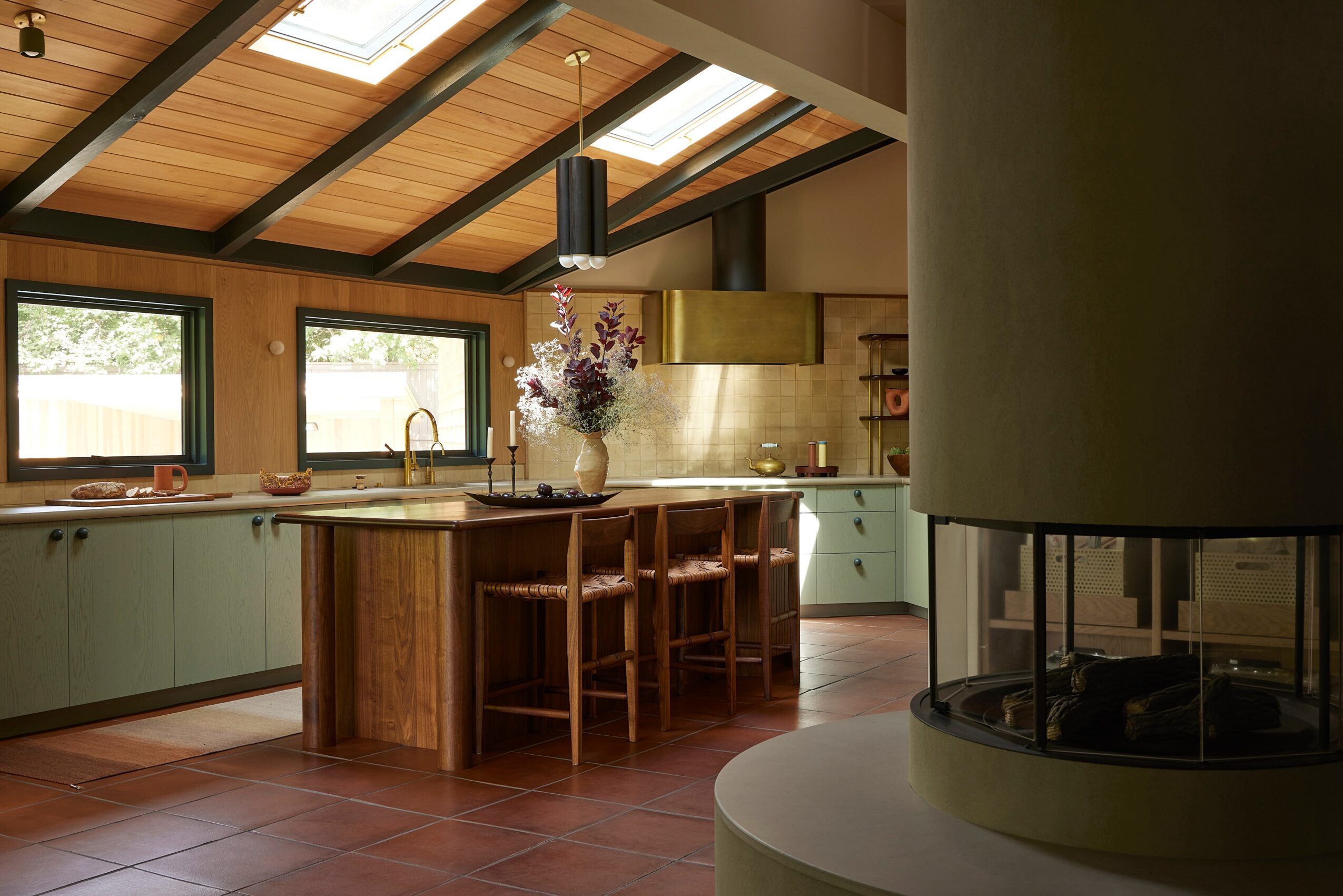 Kitchen with wooden ceiling, large windows, green cabinets, island with four chairs, and a vase with flowers, natural light.