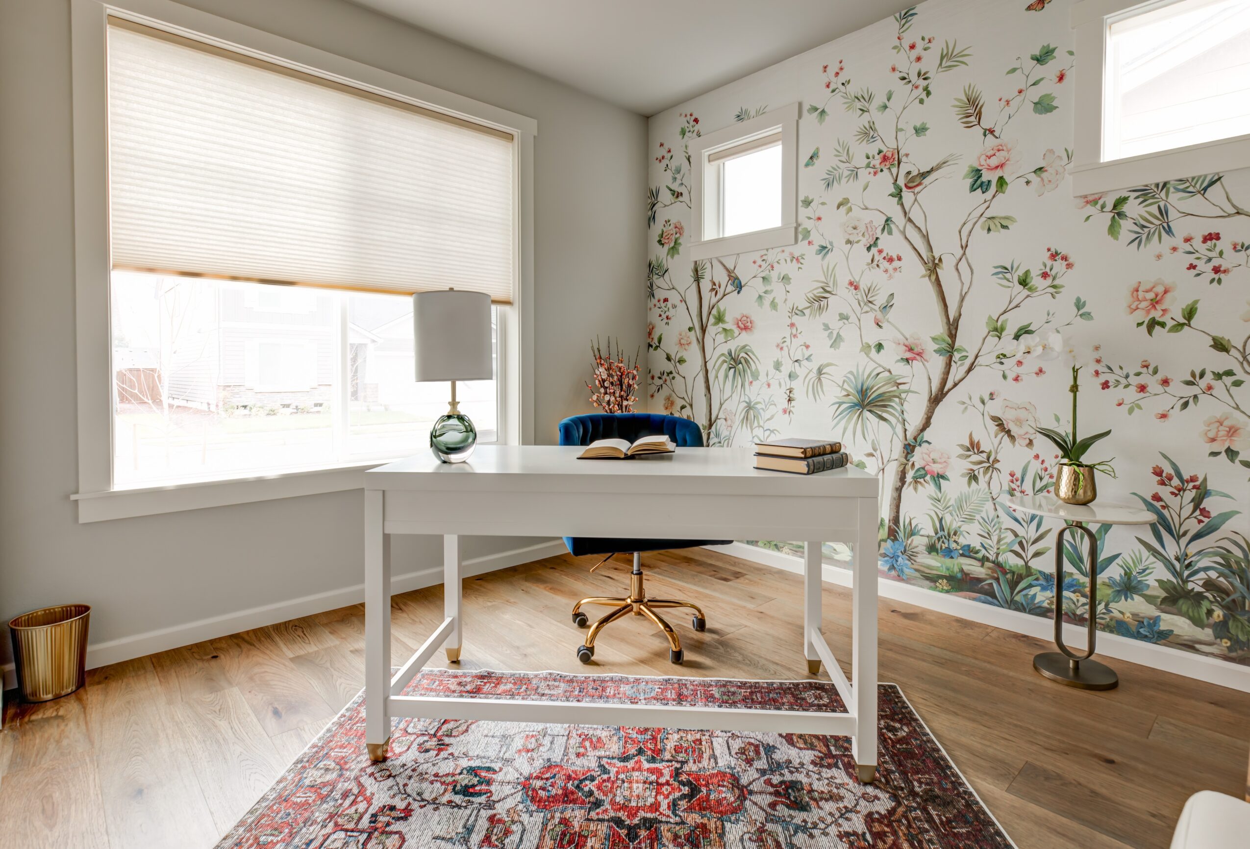 Home office with a white desk, window with blinds, floral wallpapered wall, and a small window, with a rug and decorative items.