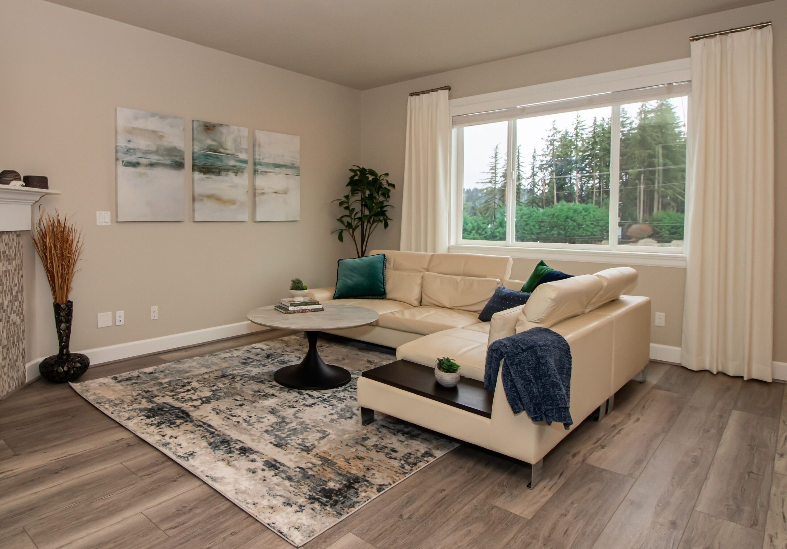 Living room with beige sofa, large window, artwork, and a patterned rug, decorated with plants and cushions.