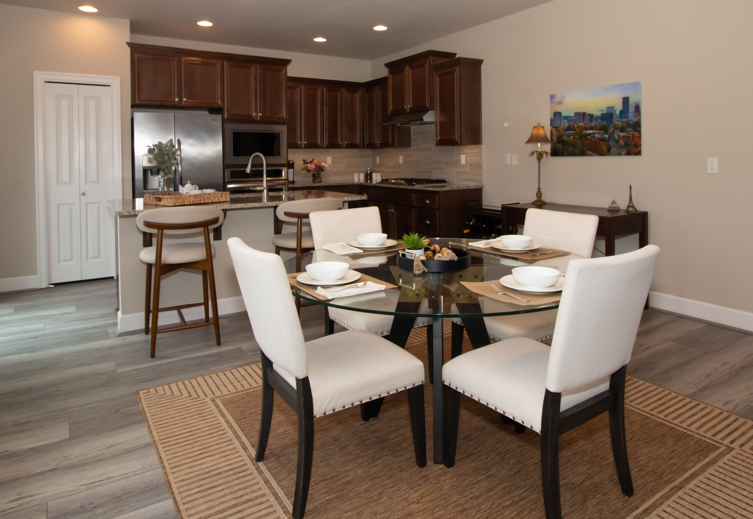 Dining area with a round glass table, six white chairs, and a kitchen with wooden cabinets in the background.