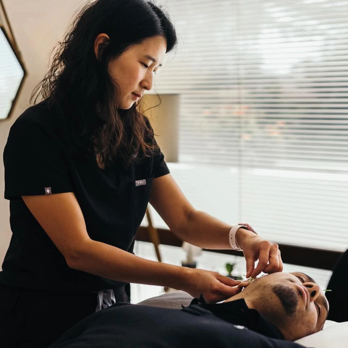A woman in black scrubs performs a procedure on a person lying down indoors with blinds in the background.
